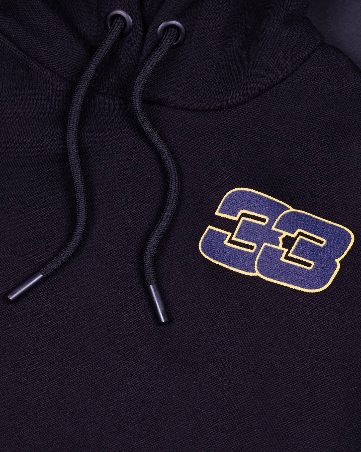 Close-up of the chest of a black hoodie with a navy 33 outlined in yellow and two black drawcords with metal tips.