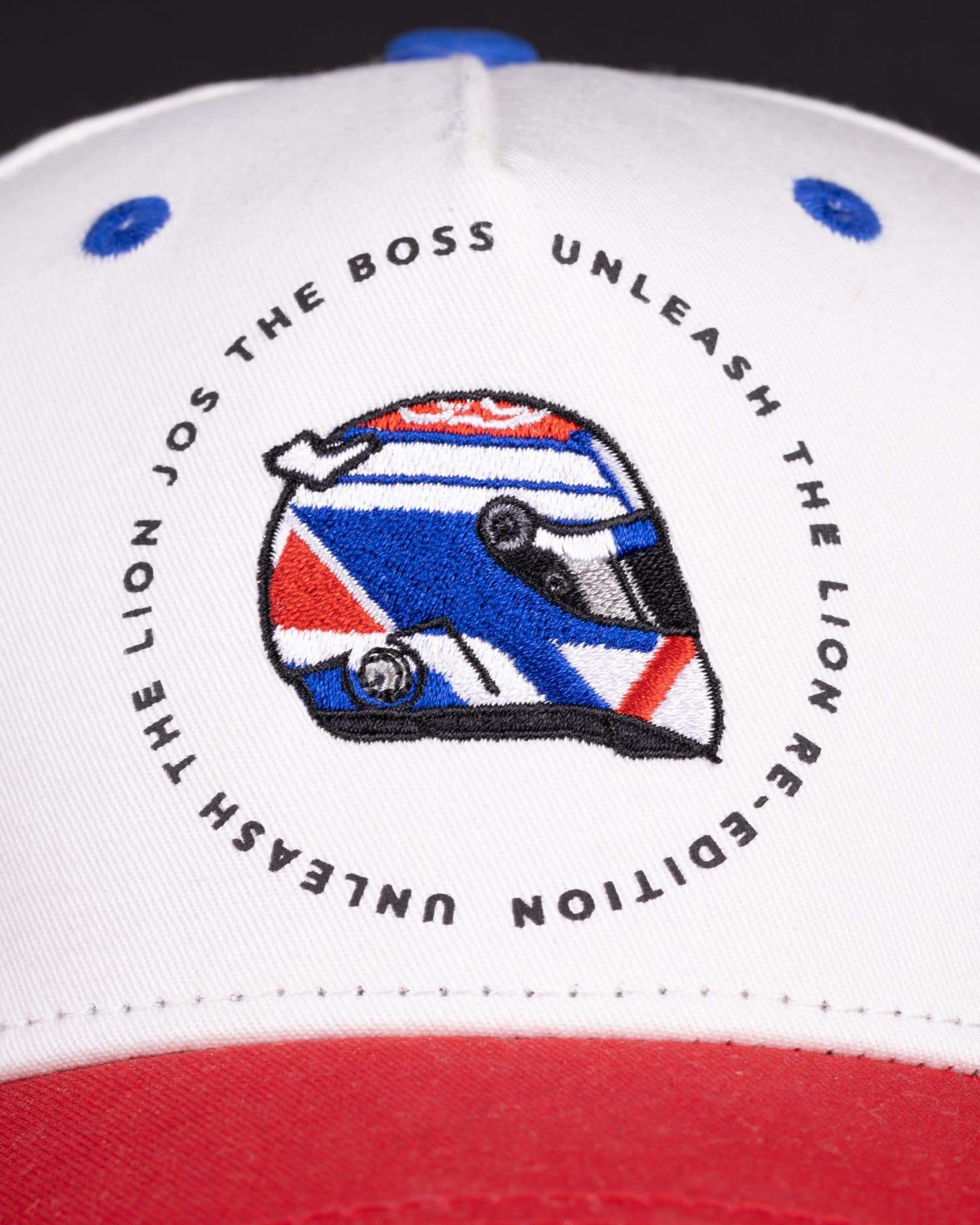 Close-up of the front of a white cap with a red peak and blue eyelets and button, embroidered with a red, white and blue racing helmet inside a ring of black lettering reading JOS THE BOSS, UNLEASH THE LION RE-EDITION and UNLEASH THE LION, against a dark background.