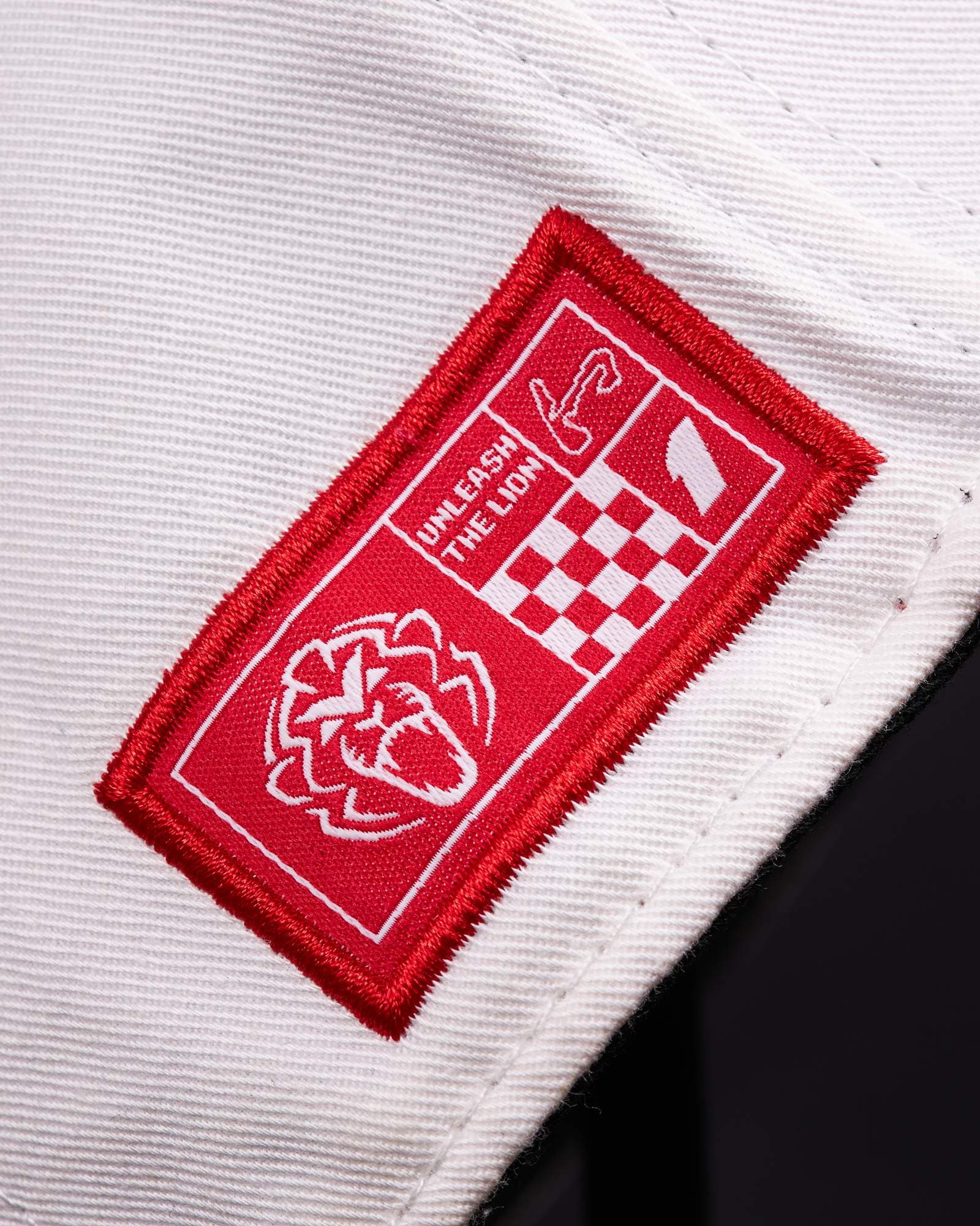 Close-up of a red woven label stitched to the white side of a cap, with a lion's head, UNLEASH THE LION, a circuit outline, a chequered pattern and a 1 in white, against a dark background.