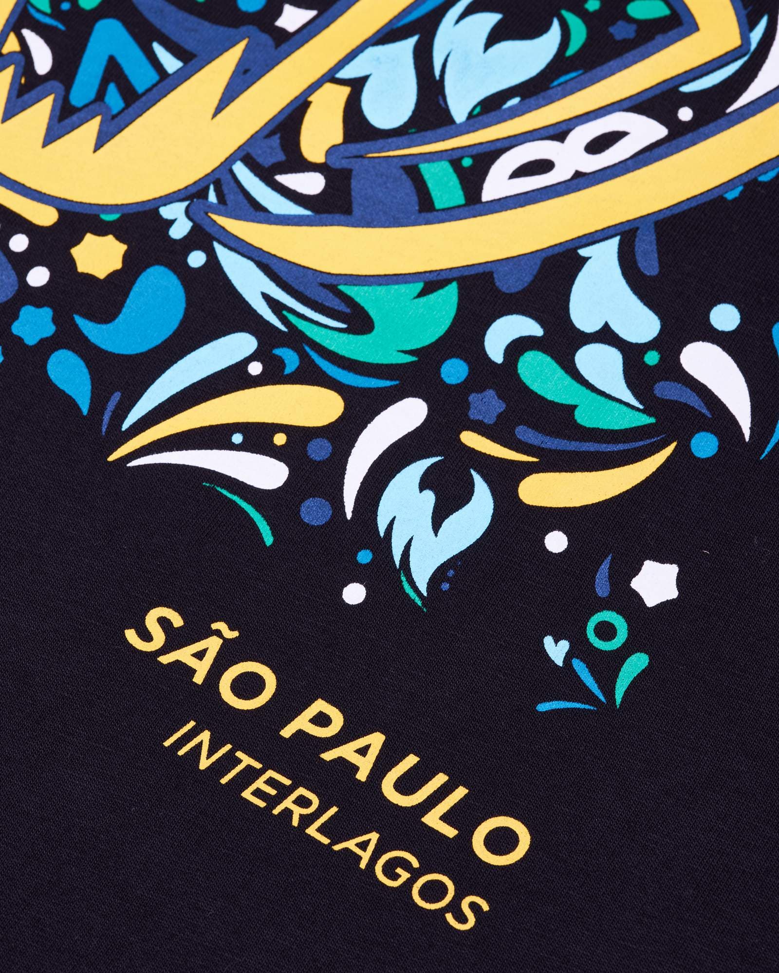 Close-up of a print on black fabric: angular pale yellow shapes edged in navy over swirls, dots, stars and small masks in blue, turquoise, green and white, above SÃO PAULO and, smaller, INTERLAGOS in yellow capitals.