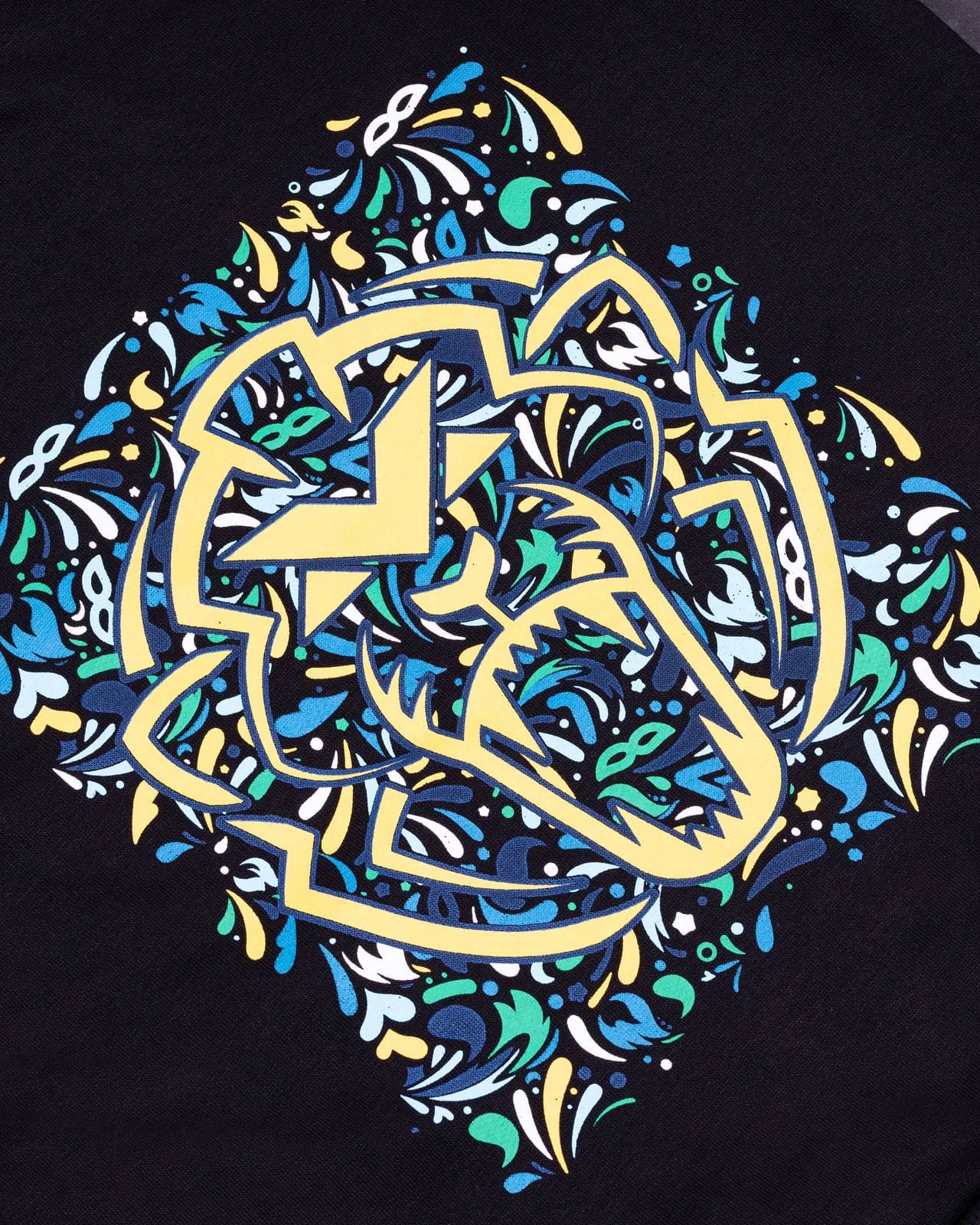 A large print on black fabric: a stylised lion's head in pale yellow outlines edged in navy, set in a diamond of blue, turquoise, green, white and yellow swirls, dots and small masks that runs off both sides of the frame.