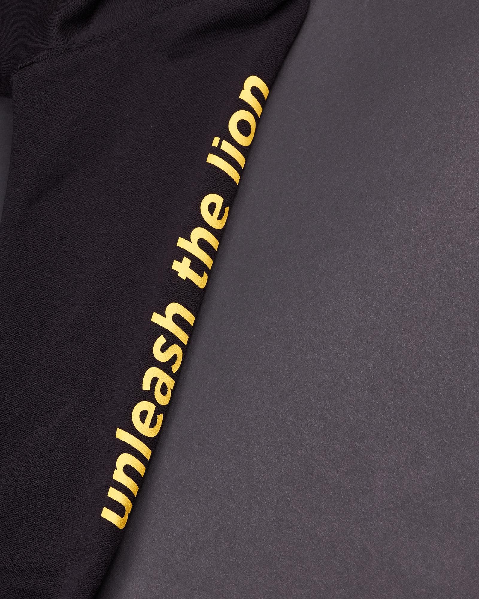 Close-up of a black sleeve printed lengthways with unleash the lion in yellow lower-case letters, on a dark grey surface.