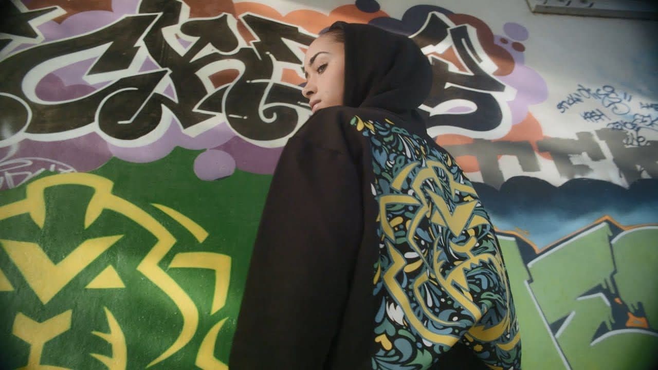 A person in a black hoodie, hood up, looking back over a shoulder in front of a graffiti wall with a yellow and green lion, the back of the hoodie printed with a lion in a yellow, blue and green carnival pattern.