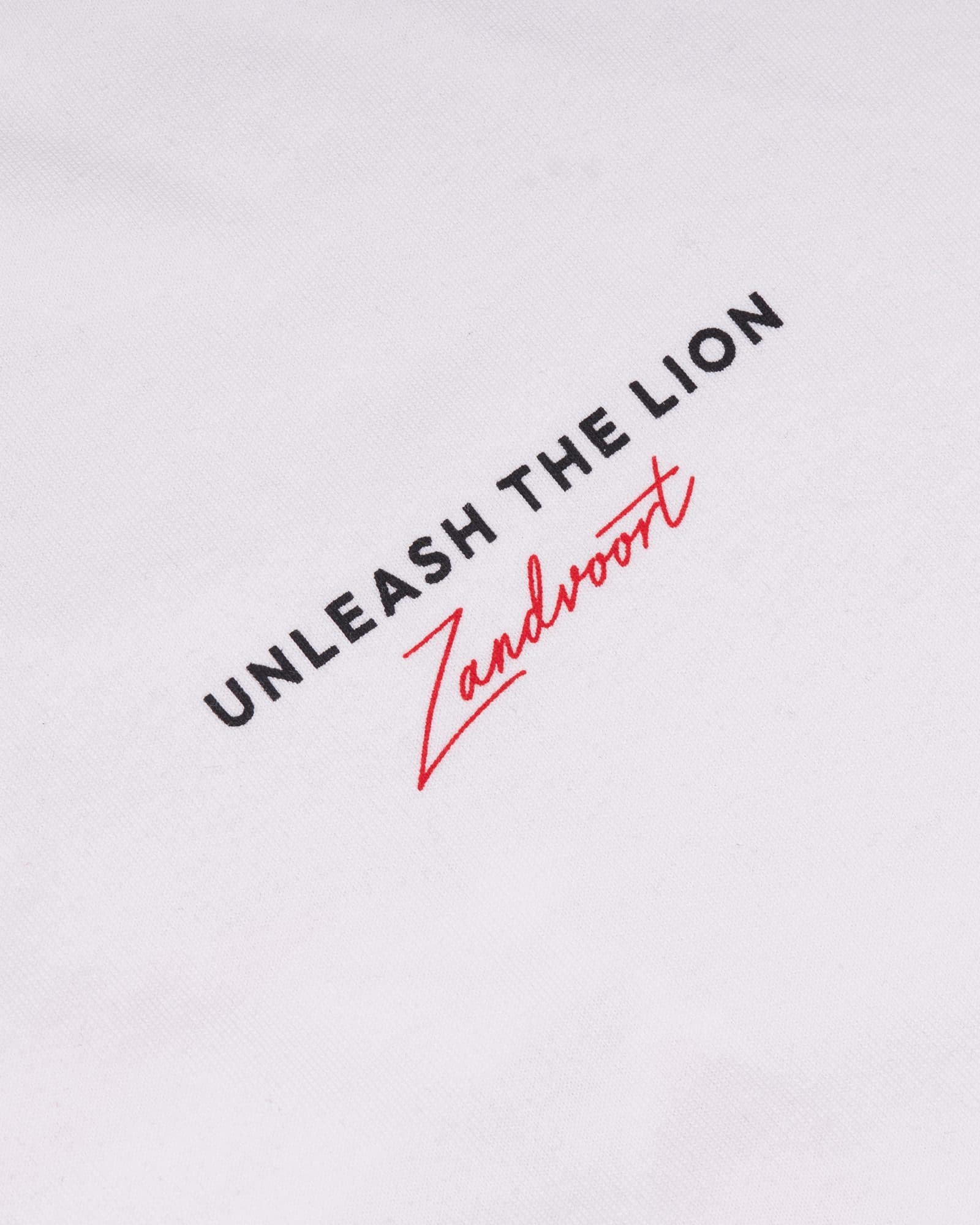 Close-up of white T-shirt fabric printed with UNLEASH THE LION in black capitals above Zandvoort in red script.