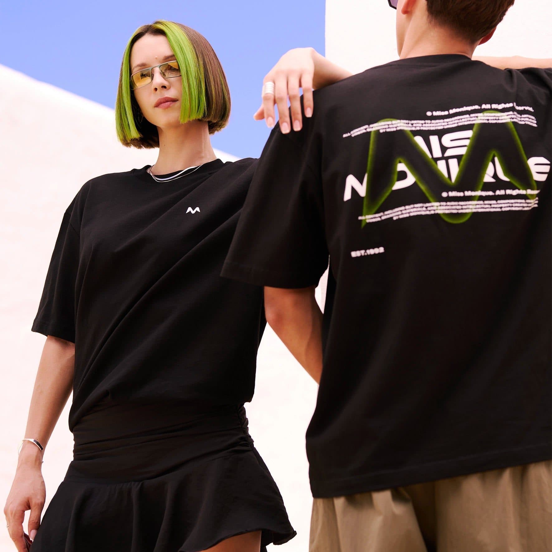 Two people in black T-shirts against a white wall and a blue sky, one with a green-streaked bob facing the camera in tinted glasses with a small white M on the chest, the other seen from behind with MISS MONIQUE in white letters behind a glowing green M, lines of small copyright text and EST.1992 on the back.