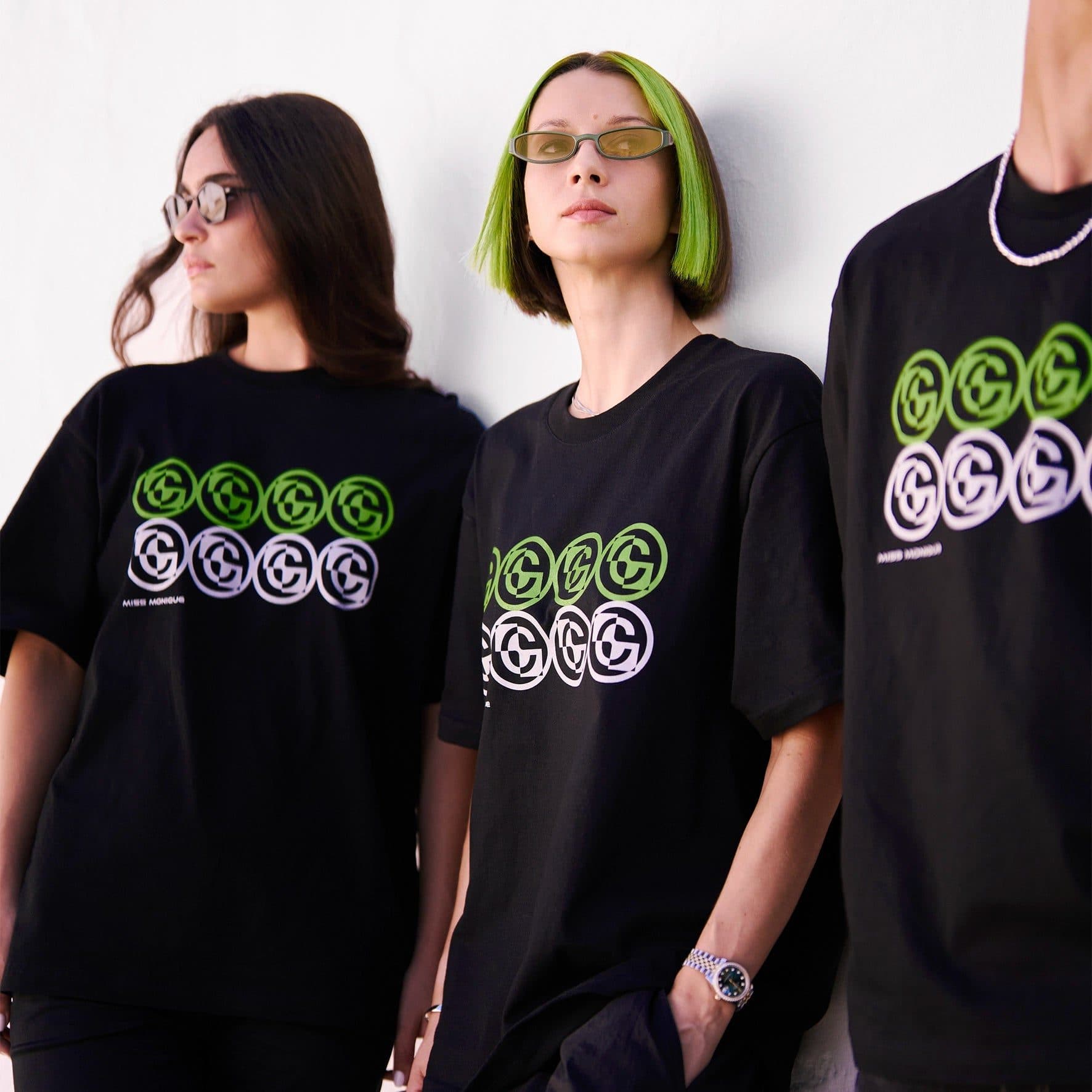 Three people in black T-shirts against a white wall, the one on the right cut off by the edge, each with a chest print of circular emblems in a green row above a white row, and MISS MONIQUE in small letters below it on two of the shirts.