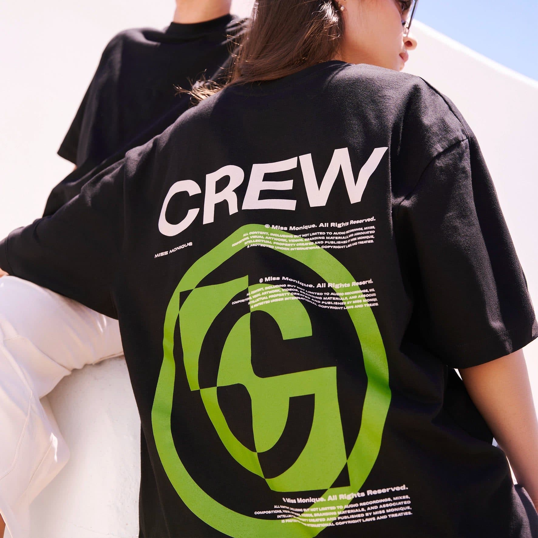 Close-up of the back of a black T-shirt in bright sun, printed with CREW in large white letters above a large green circular emblem, with MISS MONIQUE and lines of small copyright text in white, and a second person in a black T-shirt behind.