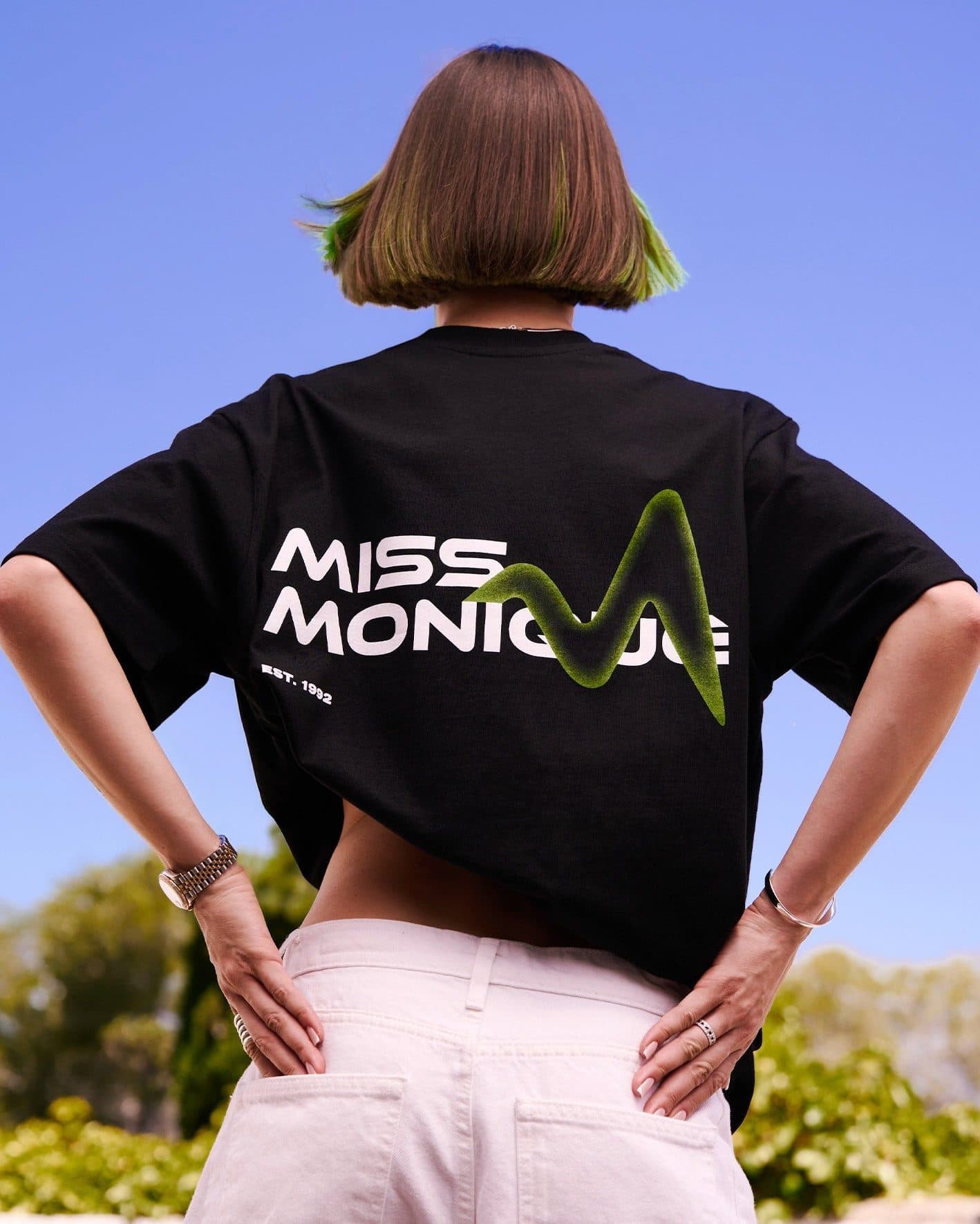 A person with a bob dyed green at the tips, seen from behind with hands on hips under a blue sky, in a black T-shirt printed on the back with MISS MONIQUE in white letters, a green wave-shaped M and EST. 1992, and white trousers.