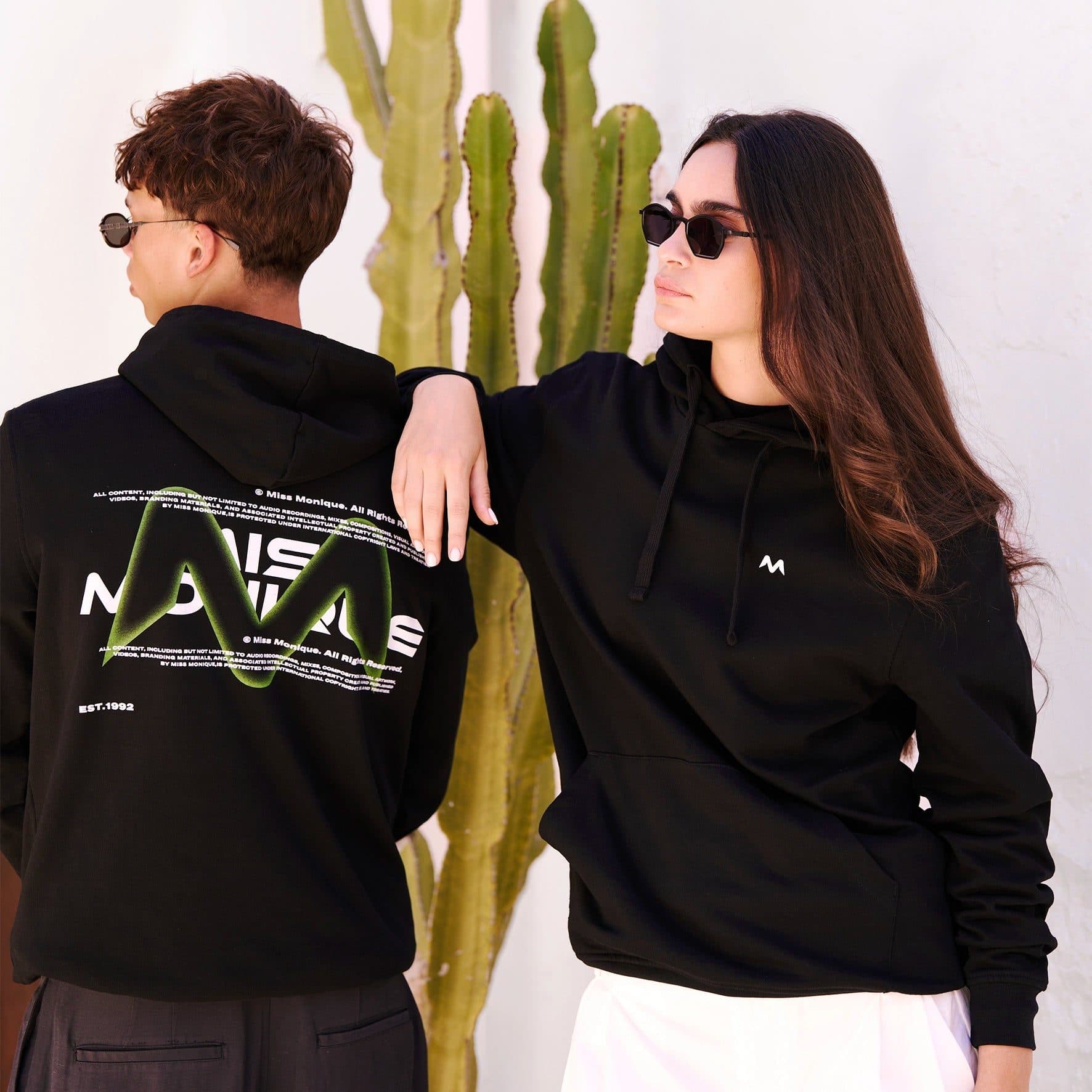 Two people in black hoodies and sunglasses against a white wall with a tall cactus between them, one seen from behind with MISS MONIQUE in white letters behind a large green M, lines of small copyright text and EST.1992 on the back, the other facing sideways with a small white M on the chest.