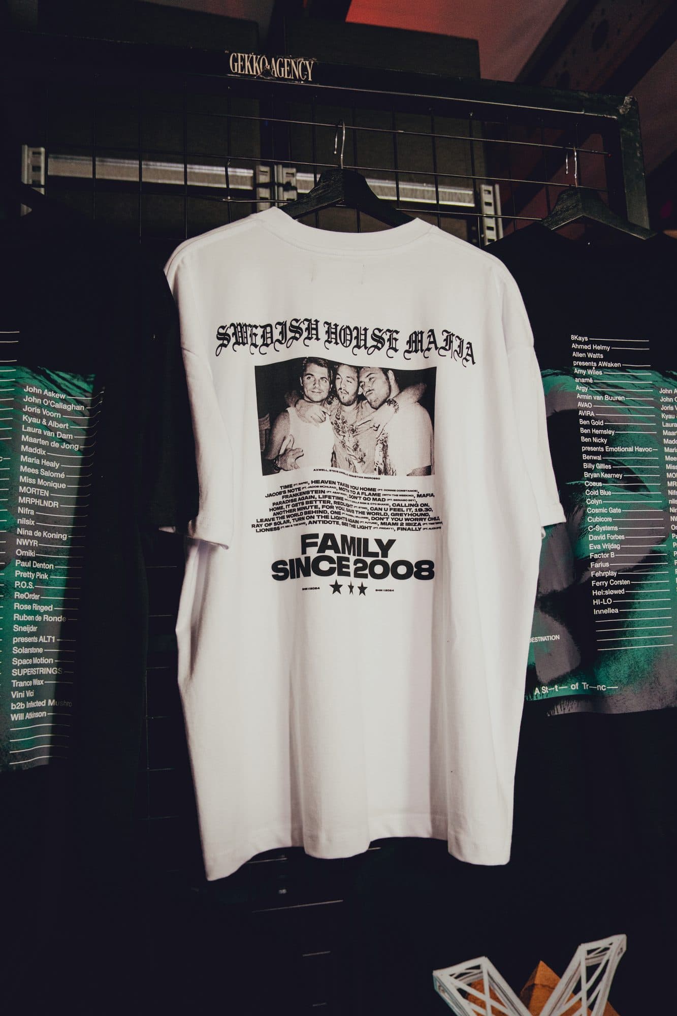 Back of a white Swedish House Mafia tee with a gothic wordmark, a black and white photo of the trio, a song list and Family Since 2008, hanging on a GEKKO Agency rack.
