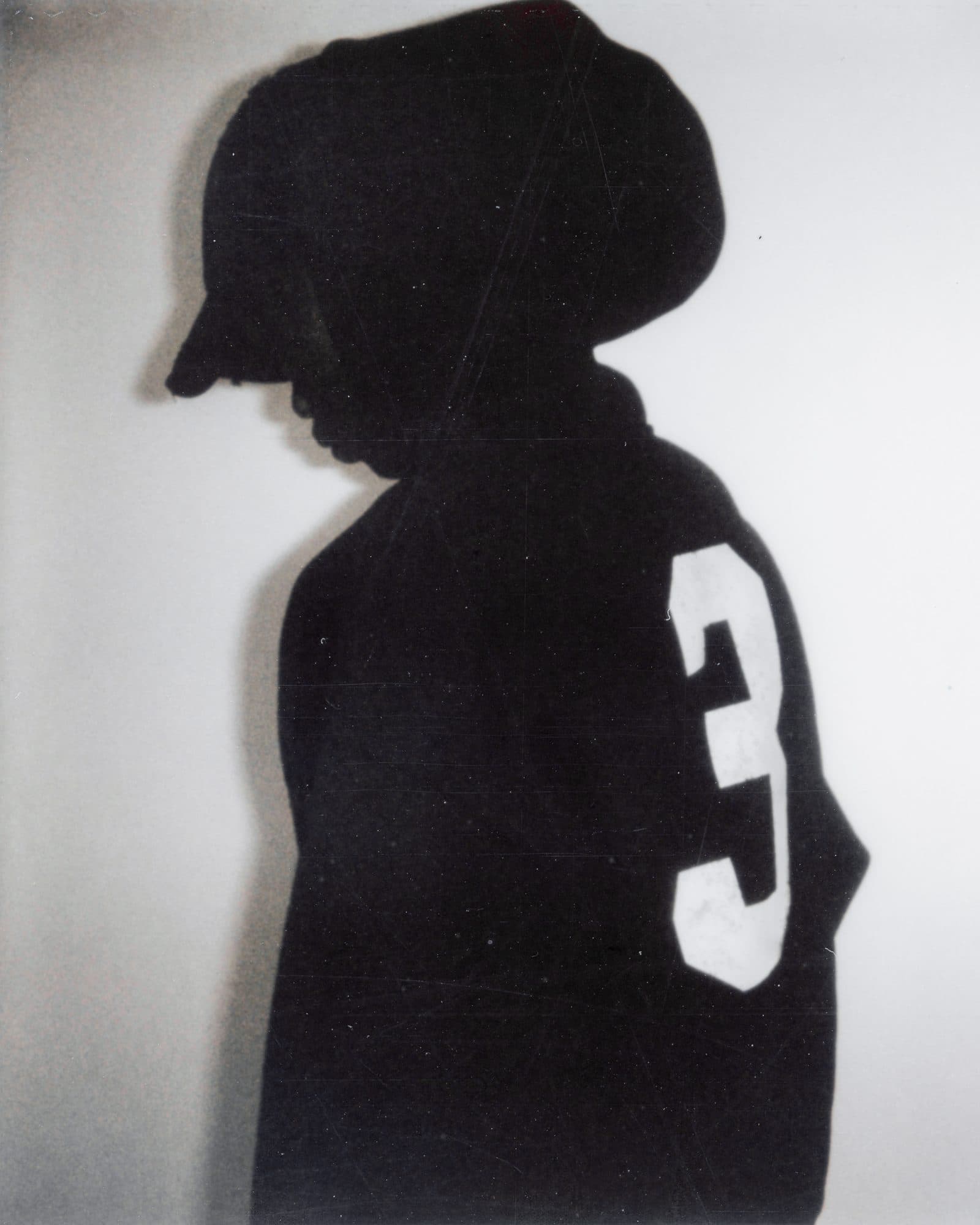 Side view of a person with head bowed in a black hoodie, hood up over a black cap, with a large white number 3 on the hoodie, against a pale wall.
