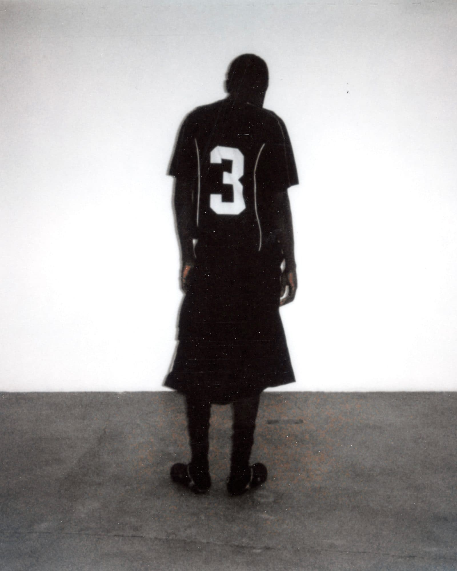 Back of a black Swedish House Mafia football shirt with a white number 3 and white piping, worn with long black shorts and black socks against a white wall.
