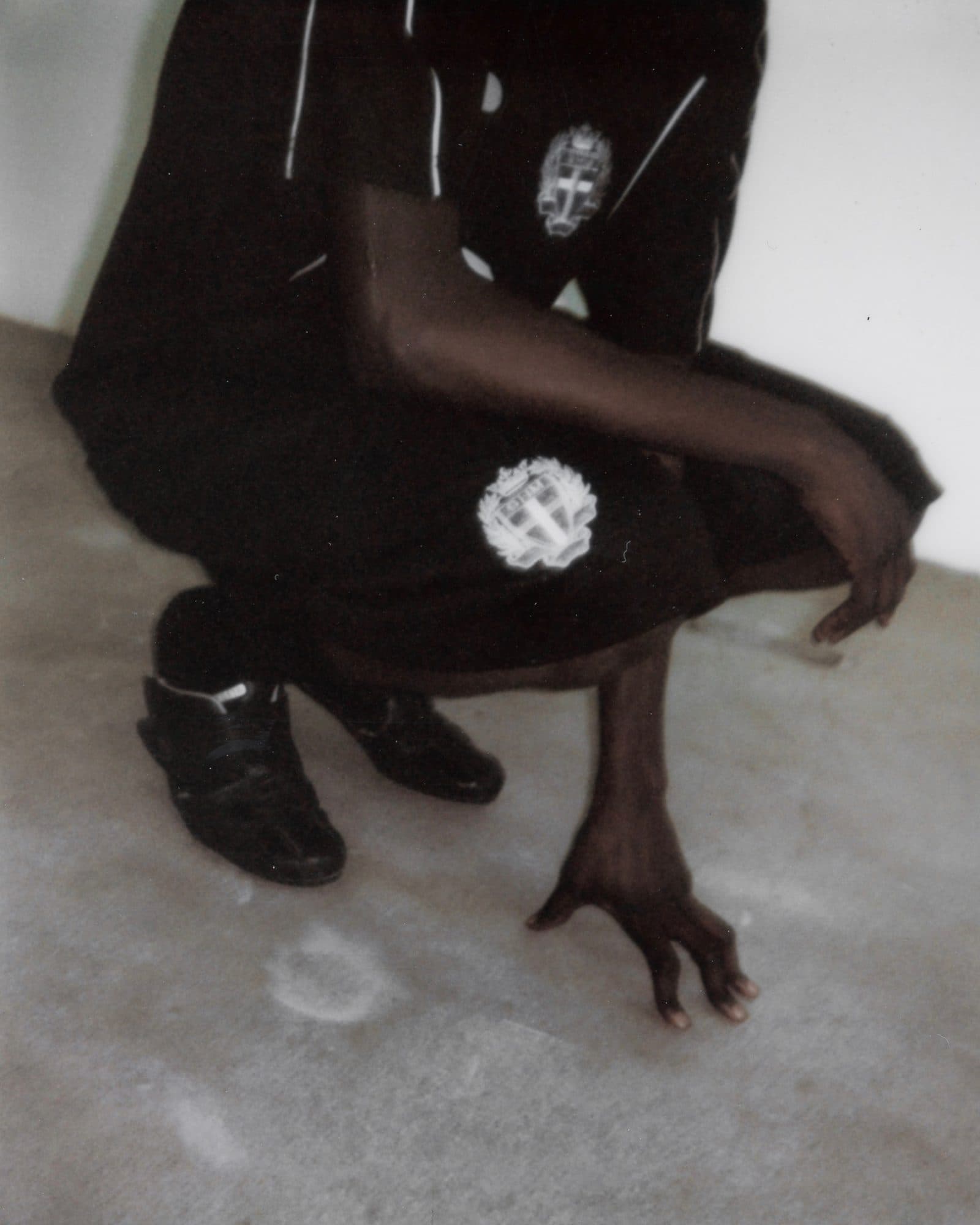 Black football shirt with white piping and a white crest on the chest, and black shorts with a large white crest on the leg, worn by a person crouching on a concrete floor in black sneakers, one hand on the floor.