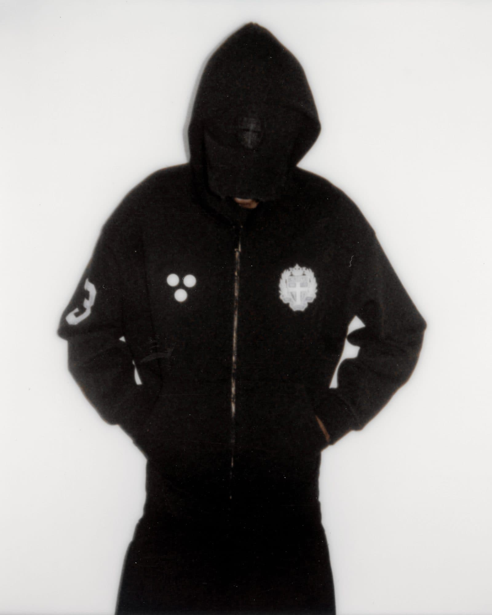 Black zip hoodie with the hood up, three white dots and a white crest on the chest and a number 3 on the sleeve, against a white wall.