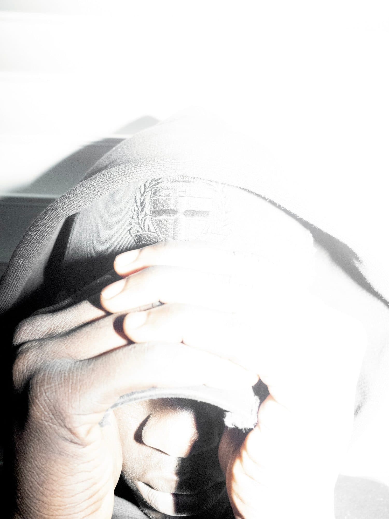 Two hands pulling a cap with a frayed brim and a tone-on-tone embroidered crest down over the eyes, the nose and mouth showing below, in hard flash light that turns the cap pale grey.