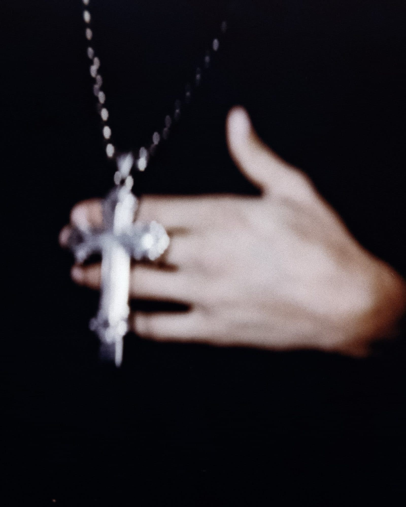 A silver cross pendant with ornate ends on a beaded chain, held against the fingers of a hand on a black ground, in soft focus.
