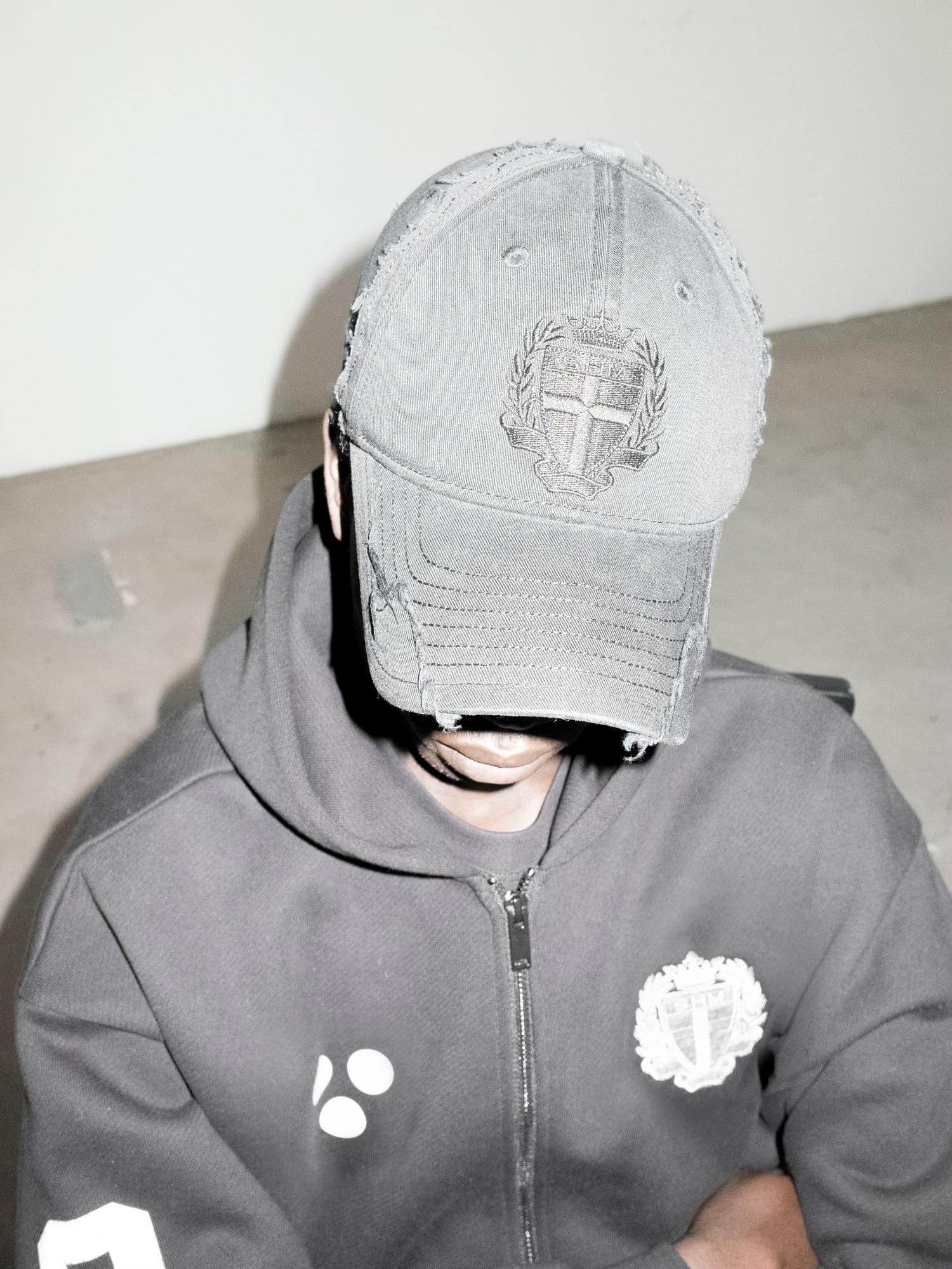 Washed black cap with frayed edges and a tone-on-tone embroidered SHM crest, worn with a black zip hoodie with a white crest and three white dots.