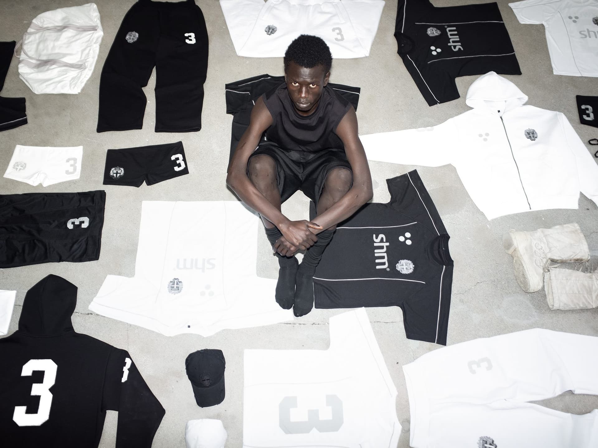A person in a black sleeveless top, black shorts and black socks sitting on a concrete floor and looking up into the camera, among black and white Swedish House Mafia football shirts with the shm wordmark, track pants, short shorts and a black cap laid out around them.