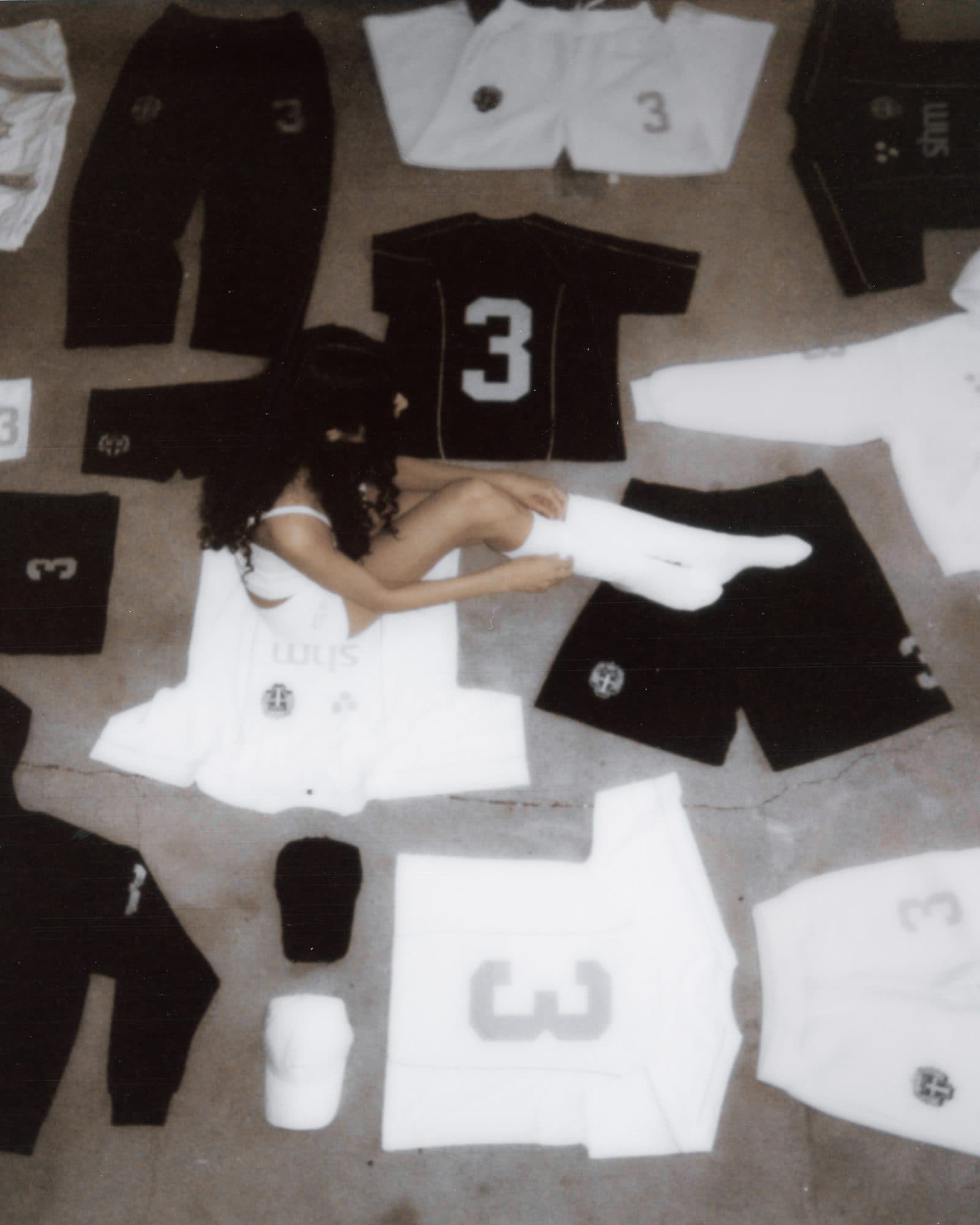 Black and white Swedish House Mafia shirts, shorts, track pants, hoodies and caps laid out on a concrete floor, with a person in white knee socks sitting on a white shm shirt.