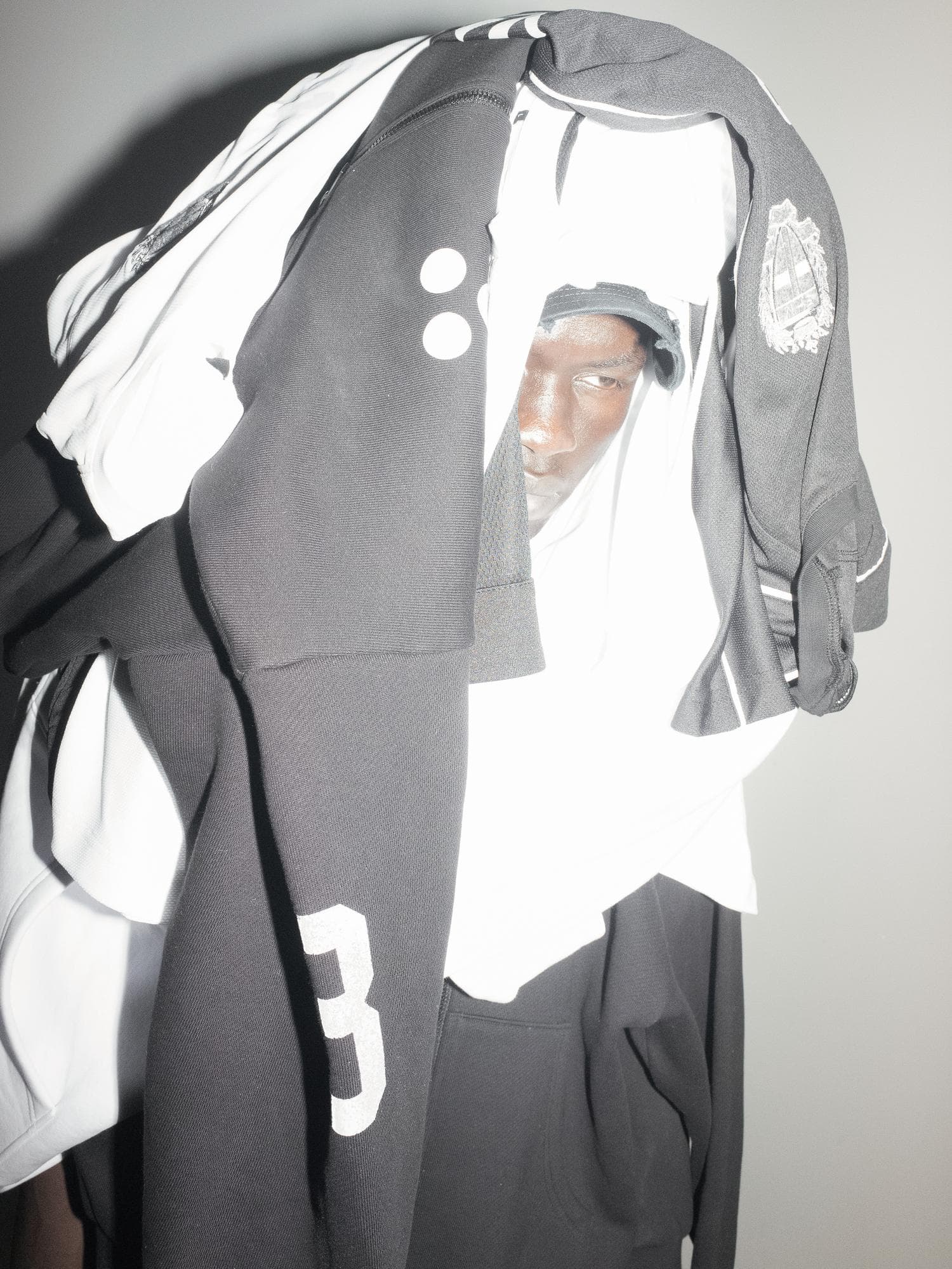 Black and white football shirts, a black zip jacket and a black hoodie piled over a person's head, one eye, the nose and the mouth showing between them, with a tone-on-tone embroidered crest on a black sleeve, white dots and a white number 3.