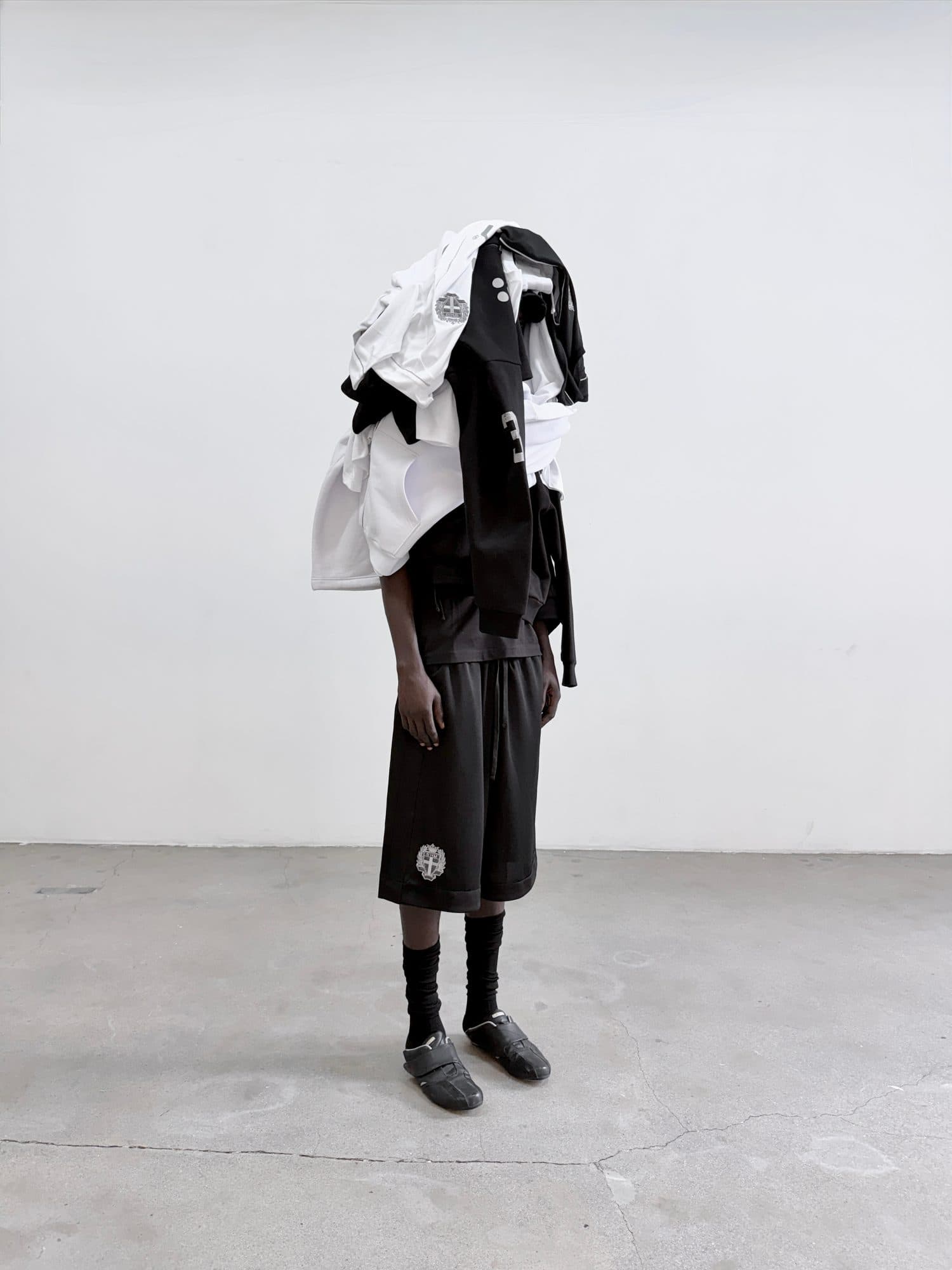 A pile of black and white Swedish House Mafia shirts, jackets and hoodies over a person's head and shoulders, worn with black shorts with a crest, black socks and black shoes on a concrete floor.