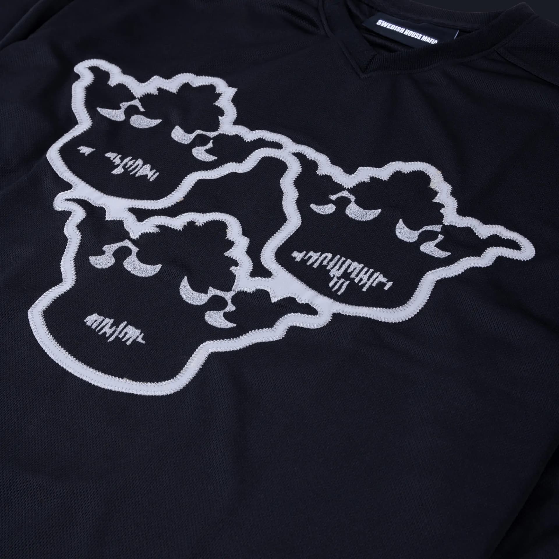 Black mesh Swedish House Mafia jersey with a white appliqué outline of three faces stitched on the chest, next to a Swedish House Mafia neck label.