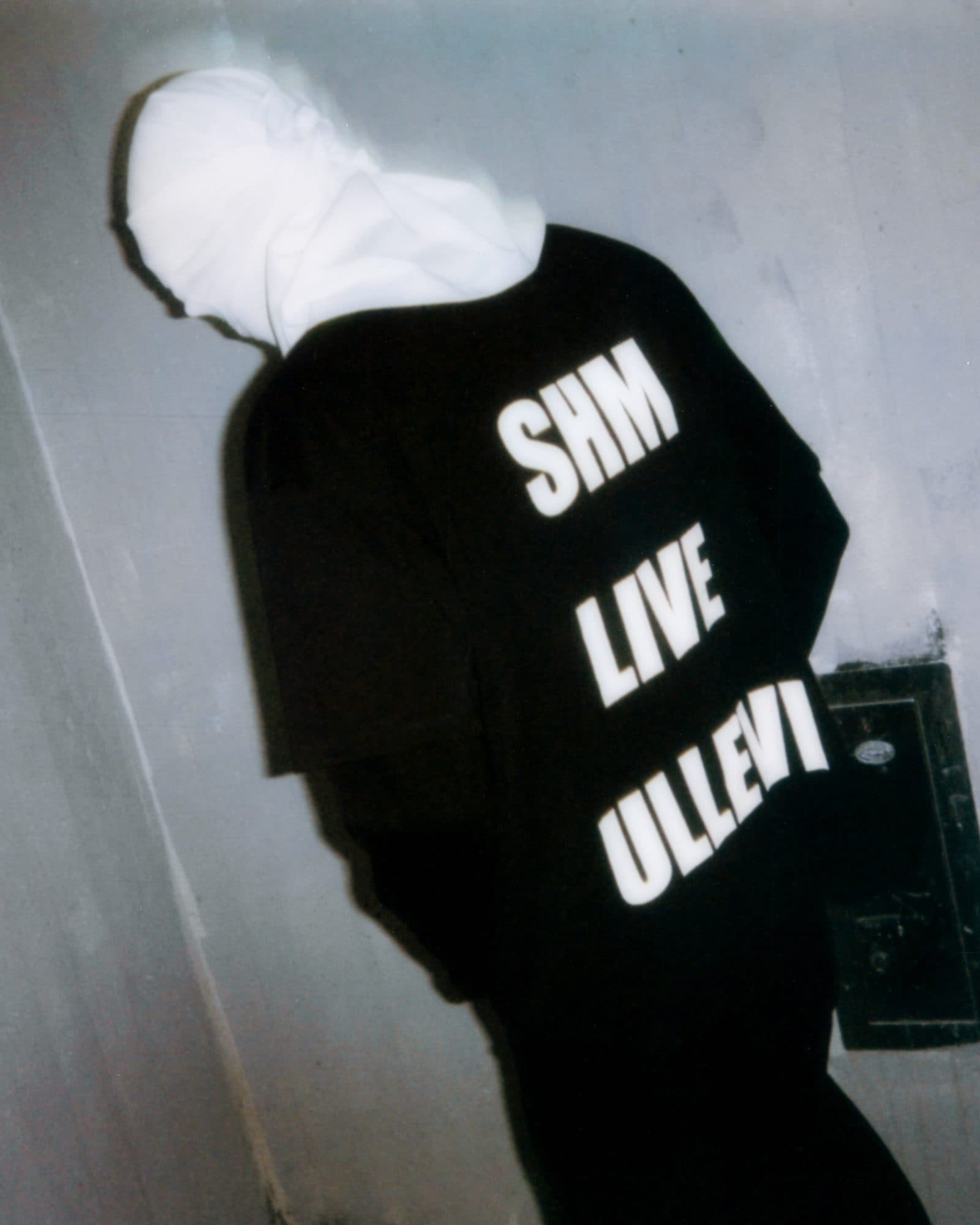 Back of a black top printed SHM LIVE ULLEVI in large white letters, worn by a person leaning against a pale wall with a white hood pulled down over the head, in flash light.