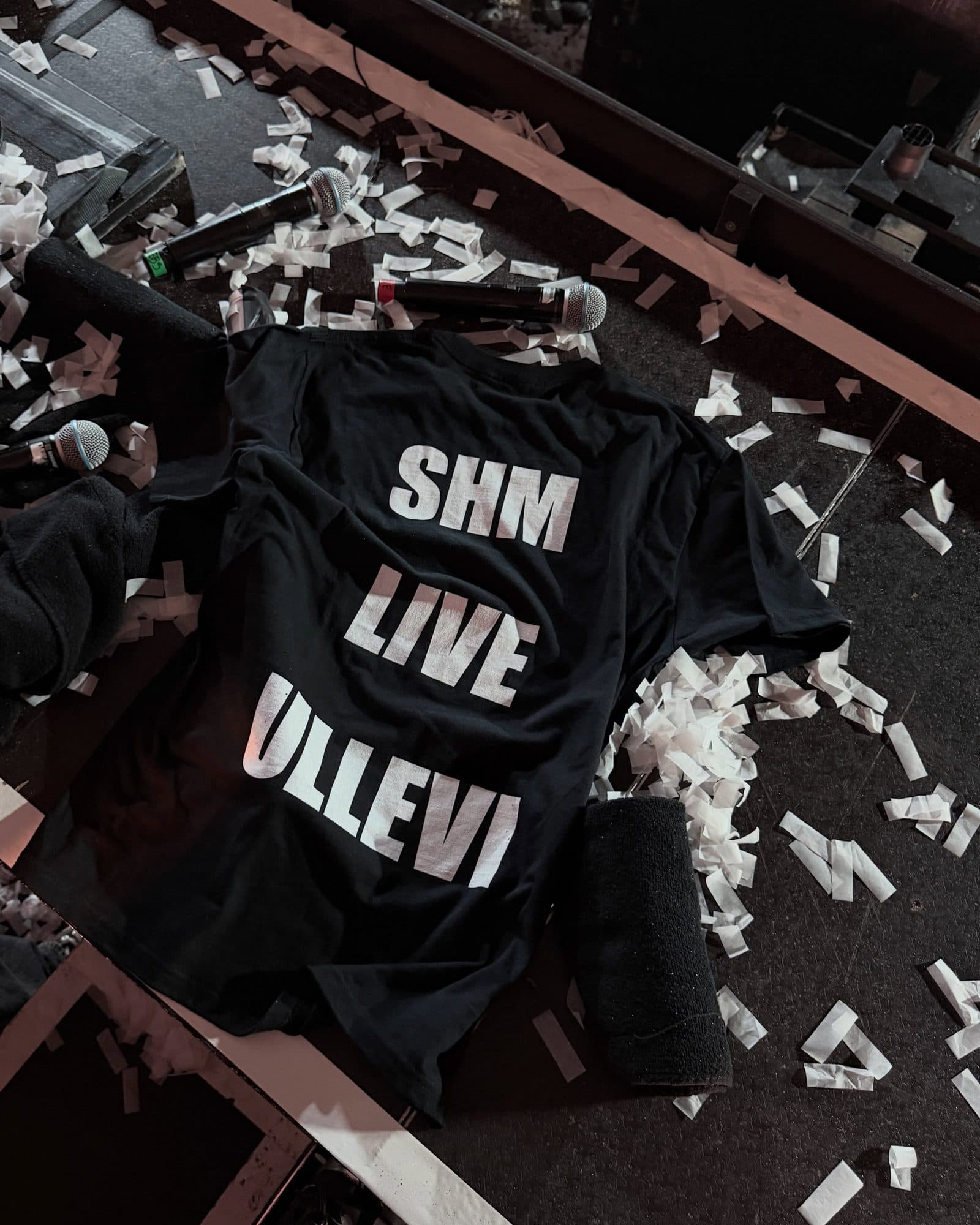 A black T-shirt printed SHM LIVE ULLEVI in large white letters, lying on a stage among white confetti, microphones and a rolled black cloth.