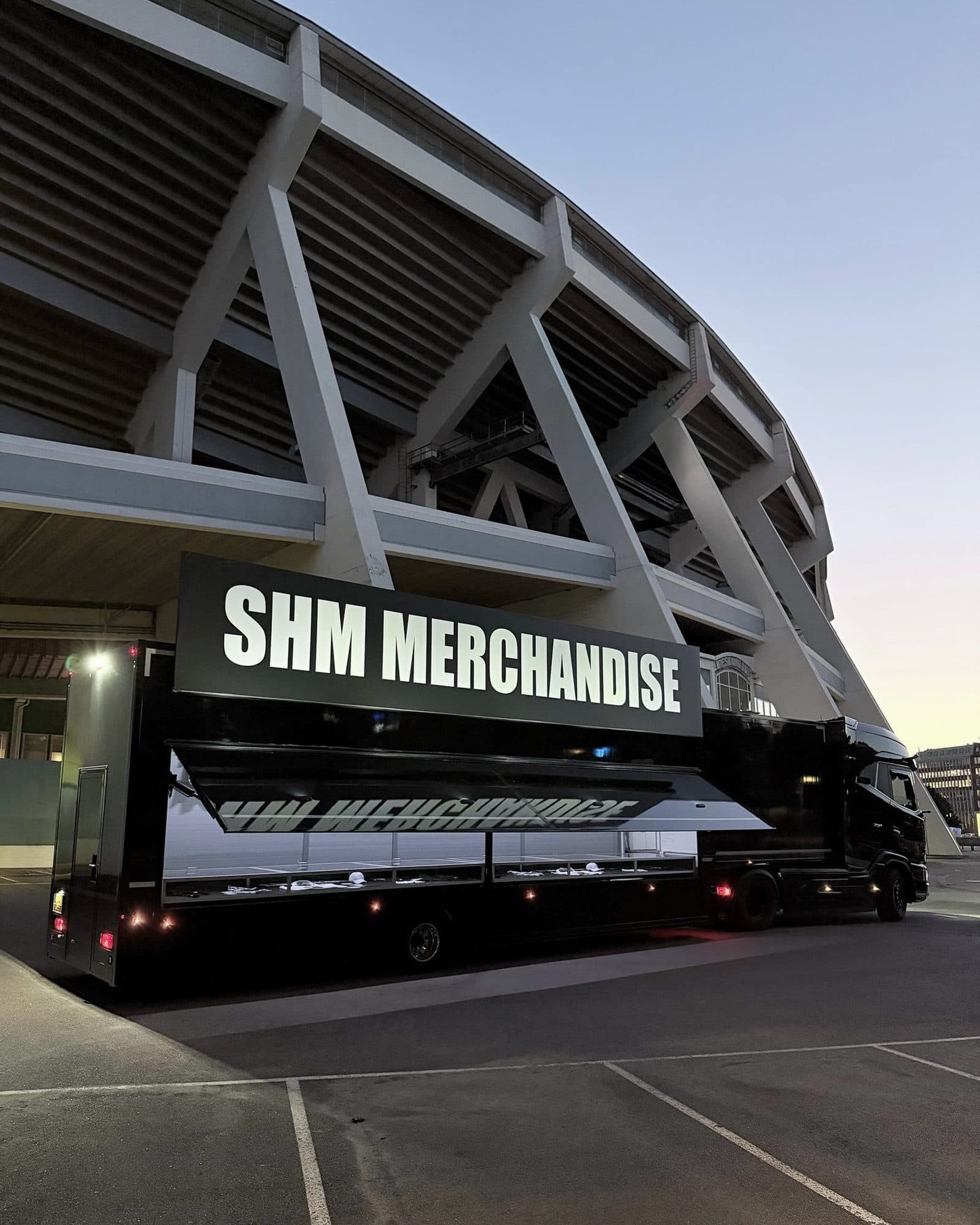 A black truck with a lit SHM MERCHANDISE sign above an open side counter, parked in front of a concrete stadium at dusk.