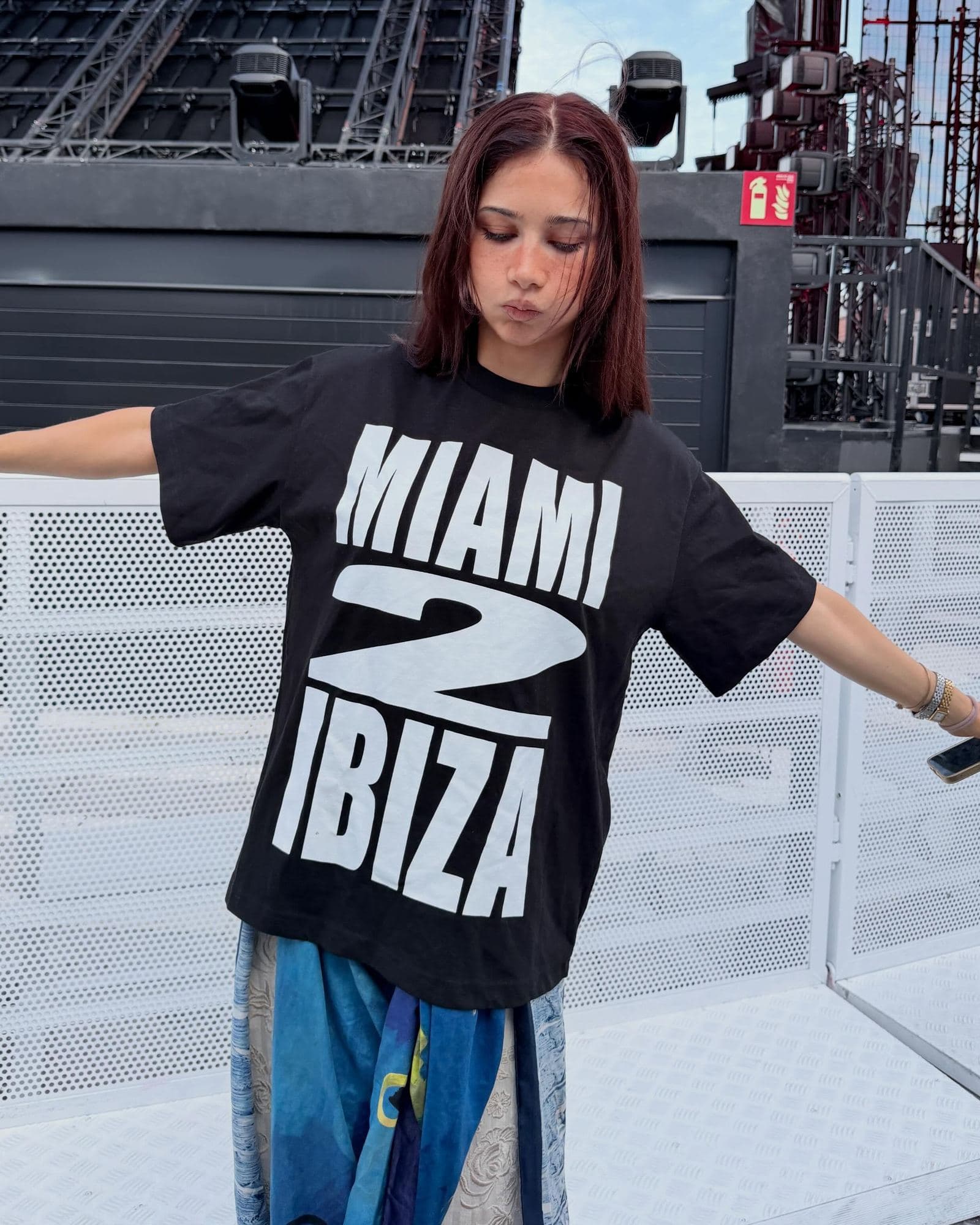A person with shoulder-length reddish-brown hair and arms spread, in a black T-shirt printed MIAMI 2 IBIZA in large white letters over a blue patterned skirt, in front of a white barrier and a stage rig with lights.