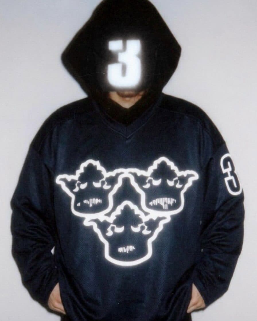 A person with a black hood up and a glowing number 3 over the face, in a dark mesh V-neck jersey with a reflective outline of three faces on the chest and a 3 on the sleeve, against a white wall in flash light.