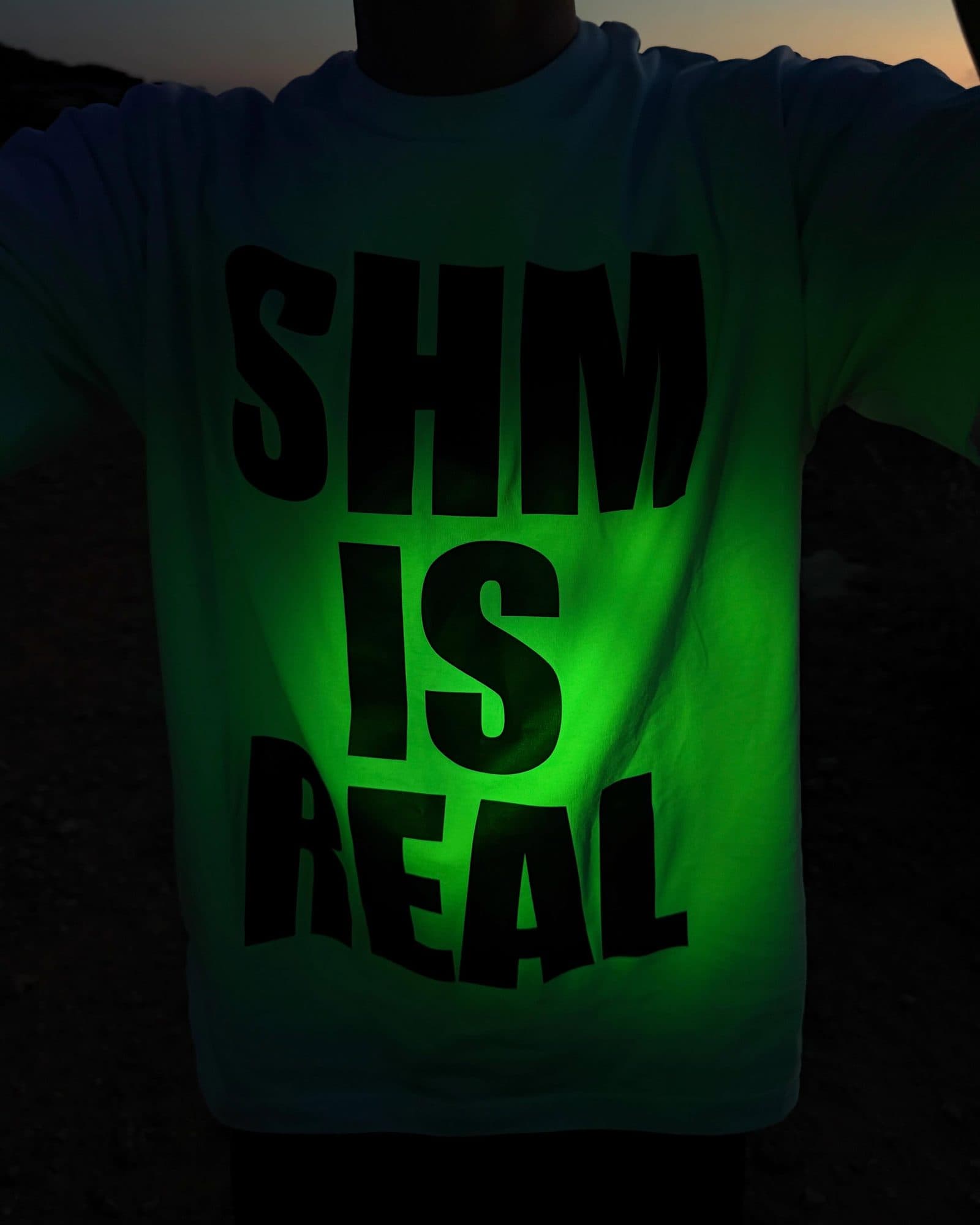A white T-shirt printed SHM IS REAL in large black letters, lit green, worn by a person with arms spread against a dark landscape at dusk.