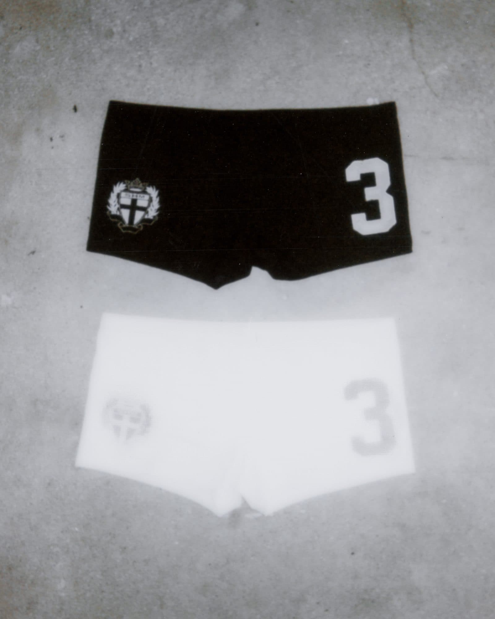 Two pairs of short shorts on a concrete floor, one black with a white SHM crest and a white 3, one white with a grey crest and a grey 3.