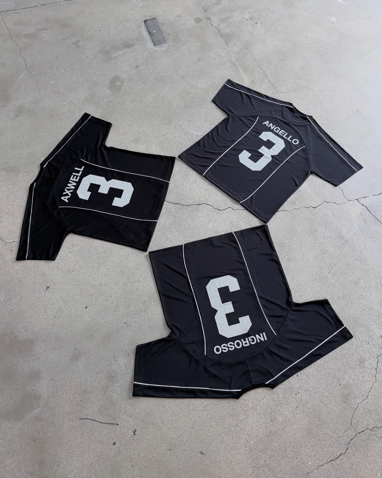 Three black football shirts with white piping laid out backs up on a cracked concrete floor, each with a white number 3 and a name: AXWELL, ANGELLO and INGROSSO.