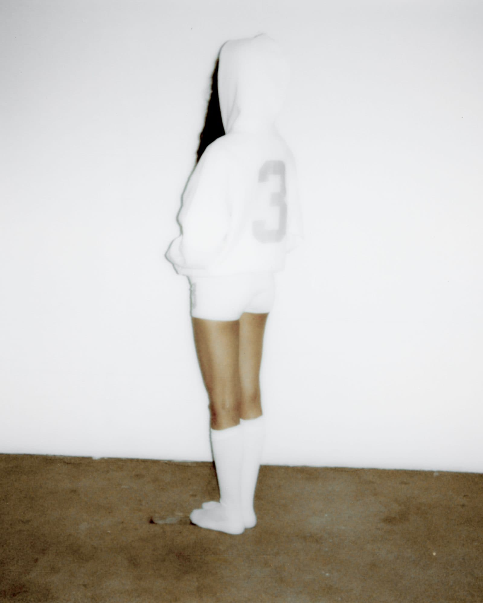 Back of a white Swedish House Mafia hoodie with the hood up and a grey number 3, worn with white shorts and white knee socks against a white wall.