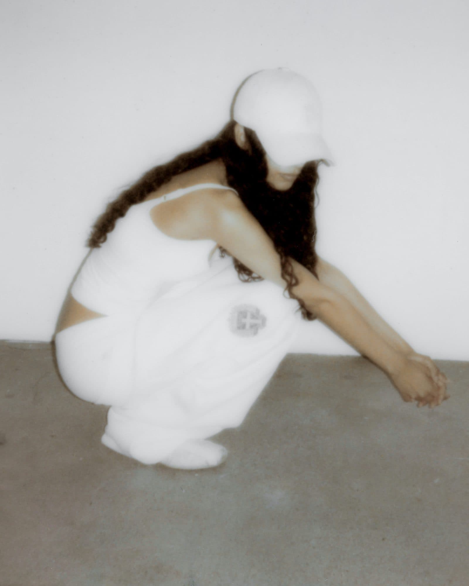 White sweatpants with a grey crest on the thigh, worn with a white strappy top, white socks and a white cap by a person with long dark curly hair, crouching on a concrete floor in front of a white wall.