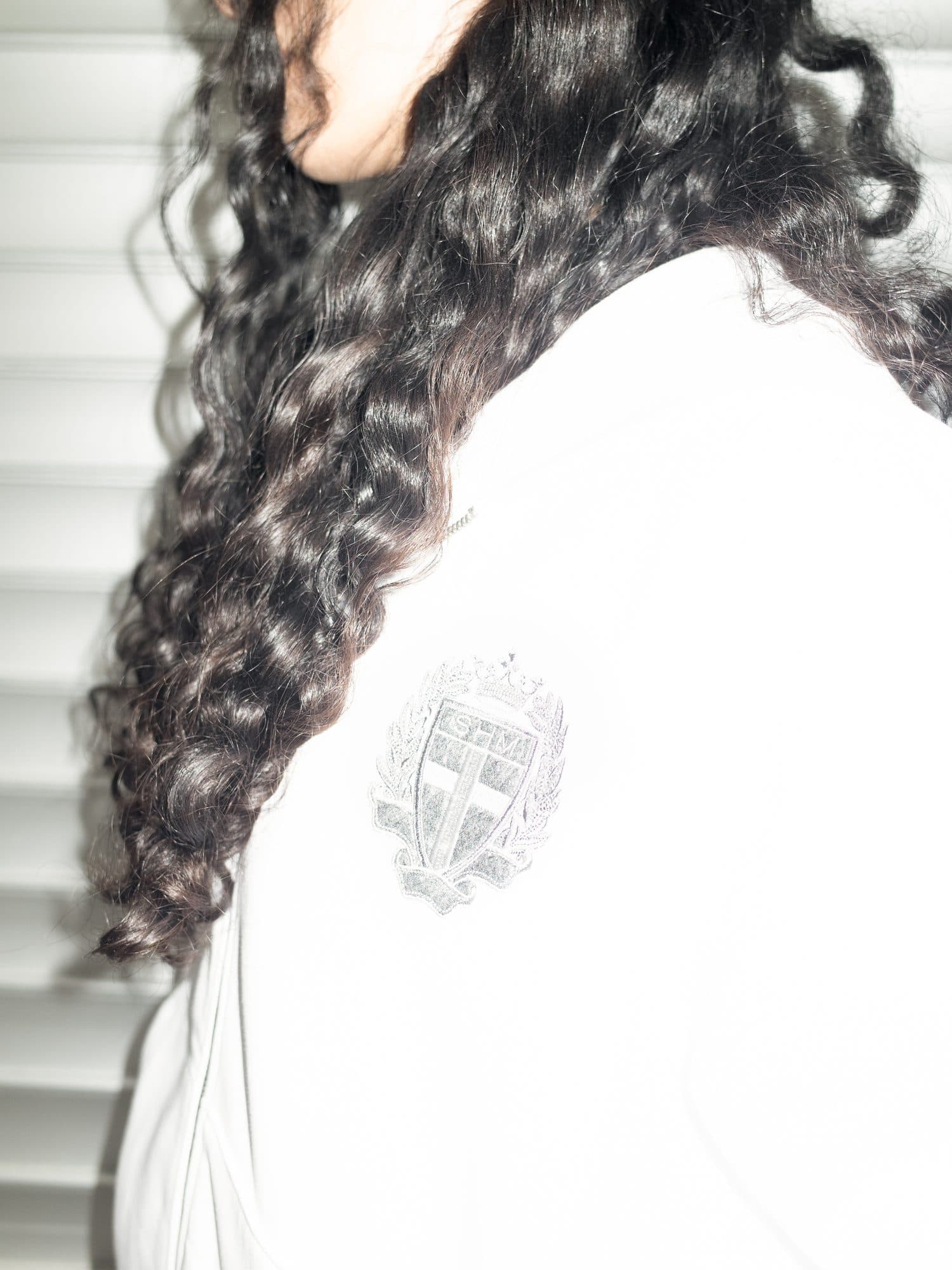 Embroidered SHM crest with a crown, laurels and a cross on the shoulder of a white top, beside long dark curly hair, in front of white blinds.