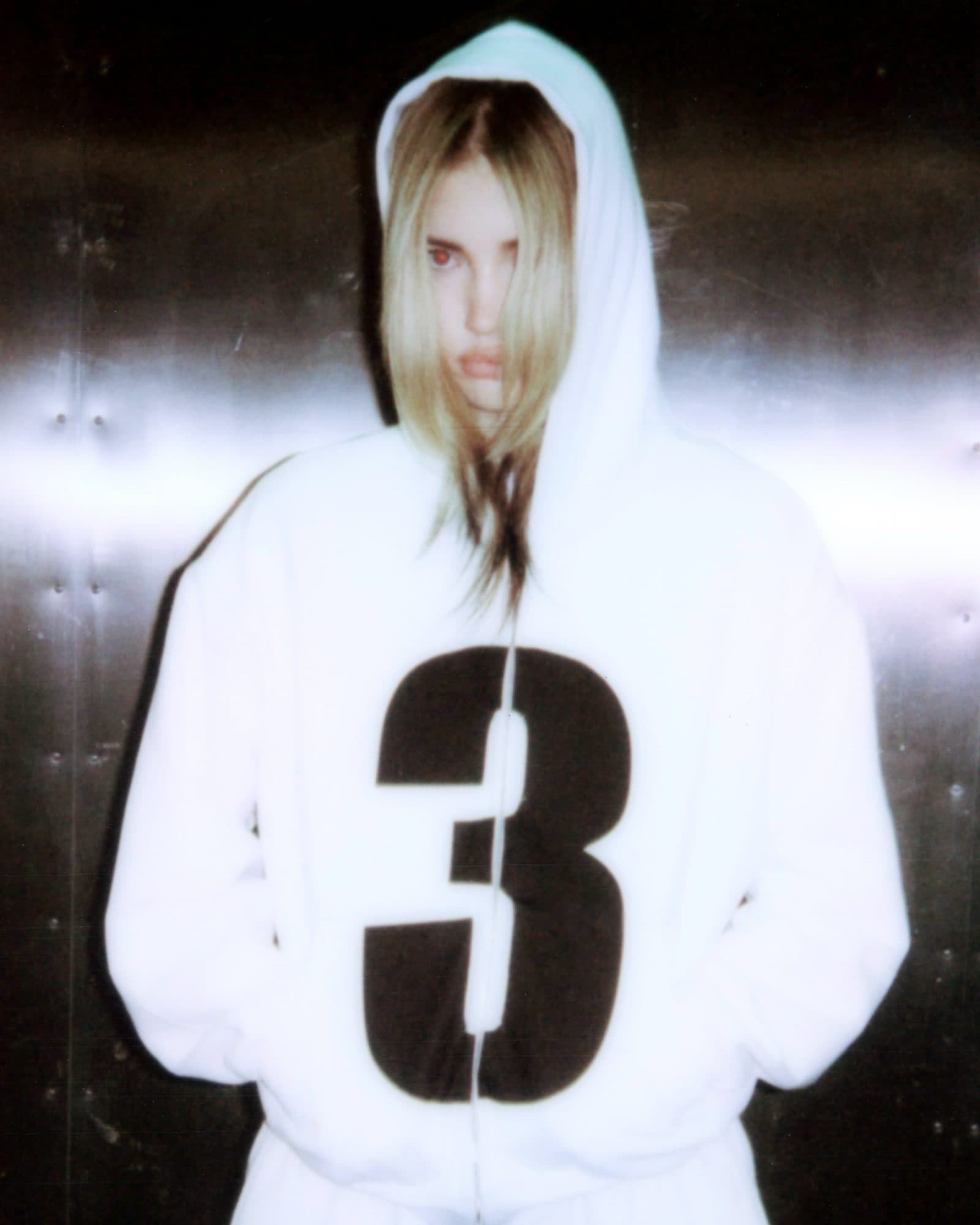 A person with long fair hair under the hood of a white zip hoodie with a large black number 3 across the front, hands in the pockets, in flash light against a steel wall.