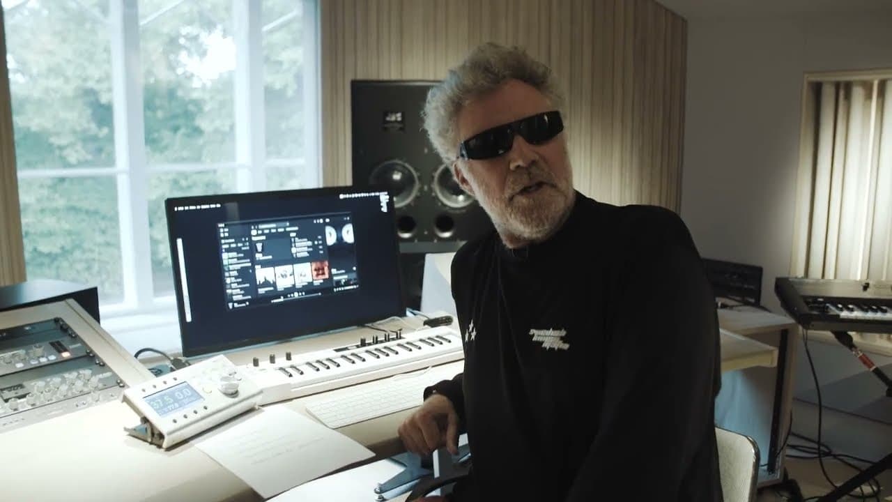 A person in black sunglasses at a desk in a music studio, in a black long-sleeved top with three small white stars and white lettering on the chest, beside a screen, a keyboard and a large studio speaker.