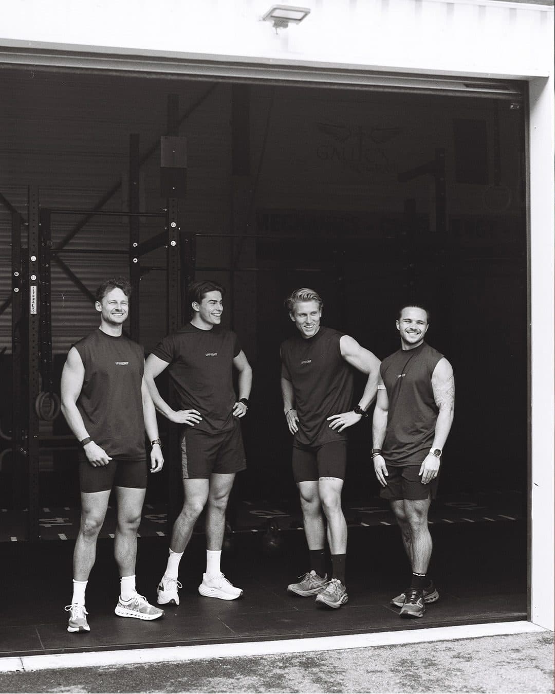 Four people in black Upfront tops with UPFRONT on the chest, three sleeveless and one with short sleeves, standing in the open door of a gym, in black and white.