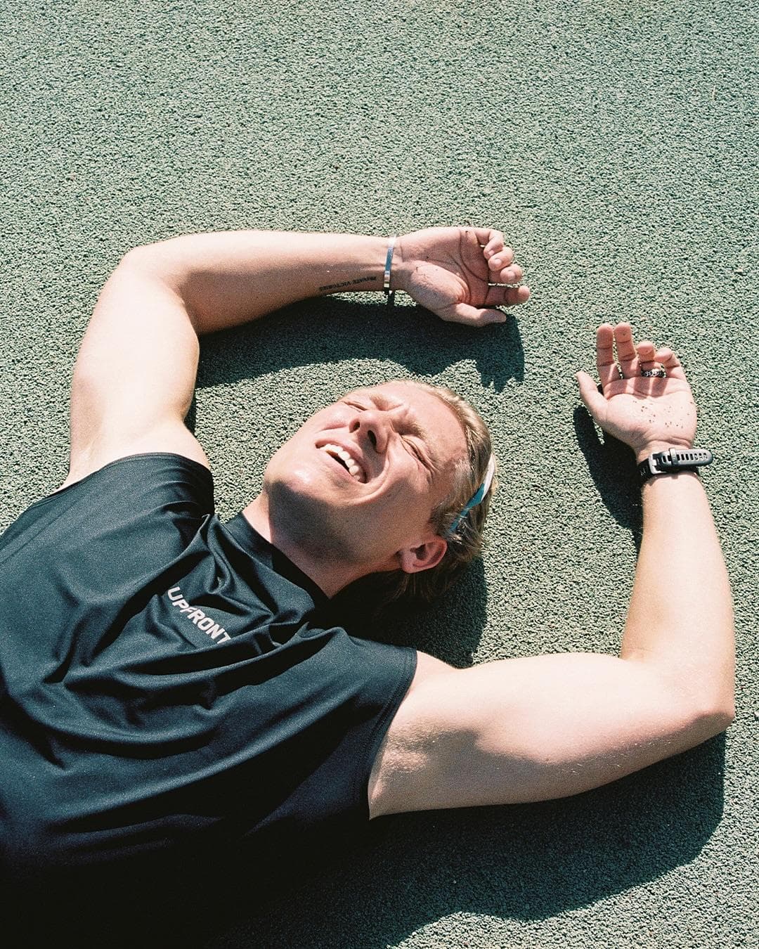 A person lying on a green outdoor sports surface with eyes closed and arms above the head, in a dark sleeveless top with UPFRONT in white letters on the chest.