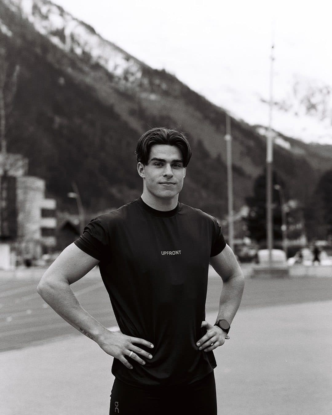 A person standing with hands on hips at an athletics track below a snowy, wooded mountainside, in a black T-shirt with UPFRONT in small white letters on the chest, in black and white.