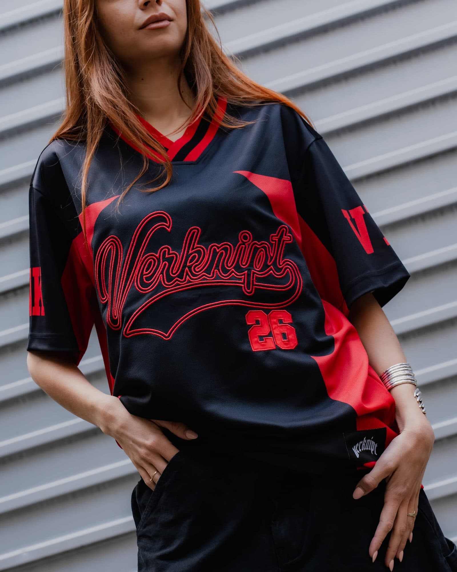A person with long red hair against a grey corrugated wall, the face cut off above the mouth by the top of the frame, in a black V-neck jersey with red side panels, a red embroidered Verknipt script and 26 on the chest, large red letters on the sleeves and a black verknipt label at the hem.