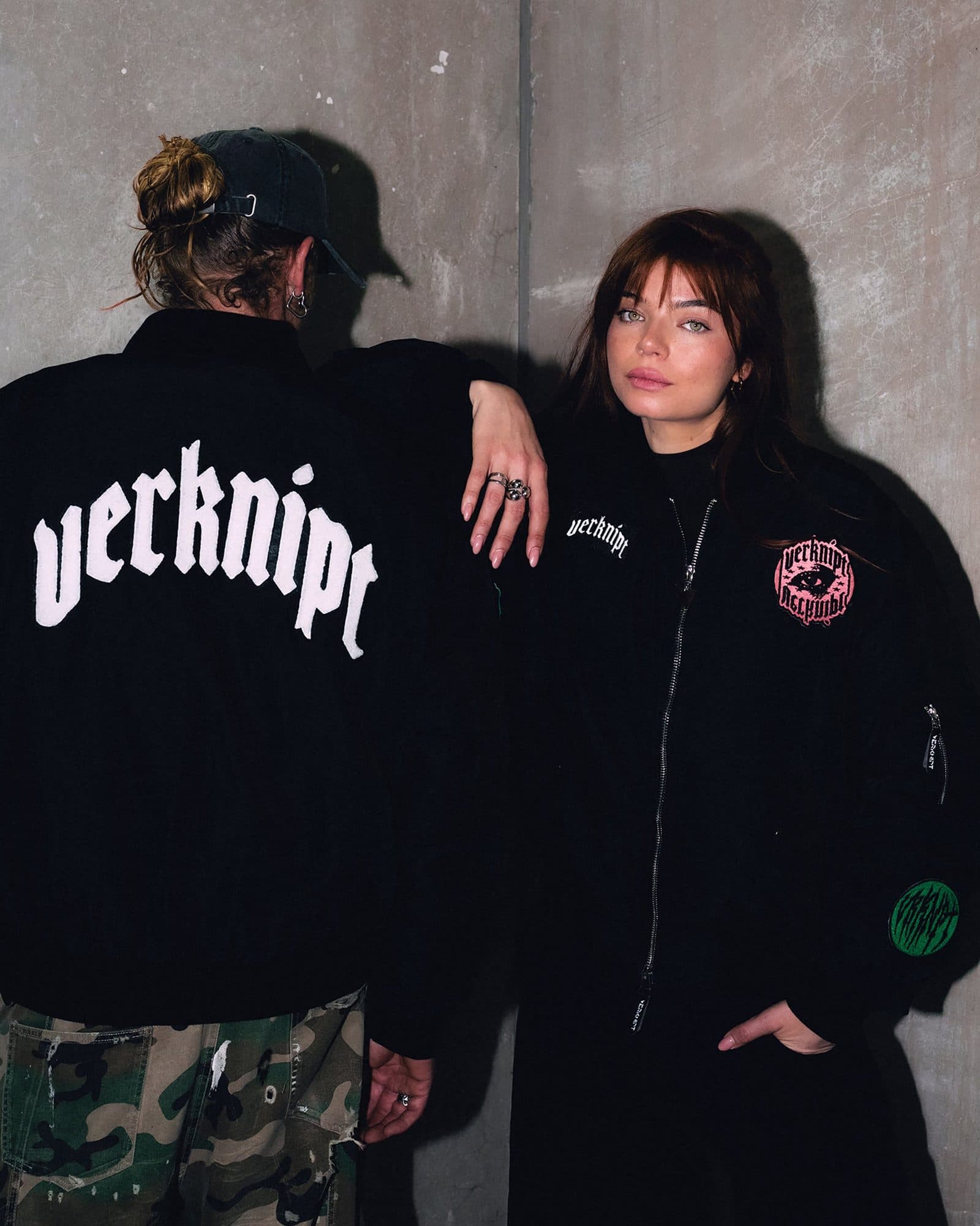 Two people against a concrete wall in black bomber jackets, one seen from behind, in a black cap and camouflage trousers, with large white verknipt lettering across the back of the jacket, the other facing the camera with a small verknipt badge on the chest, a round pink patch with an eye and verknipt lettering, and a round green patch on the sleeve.