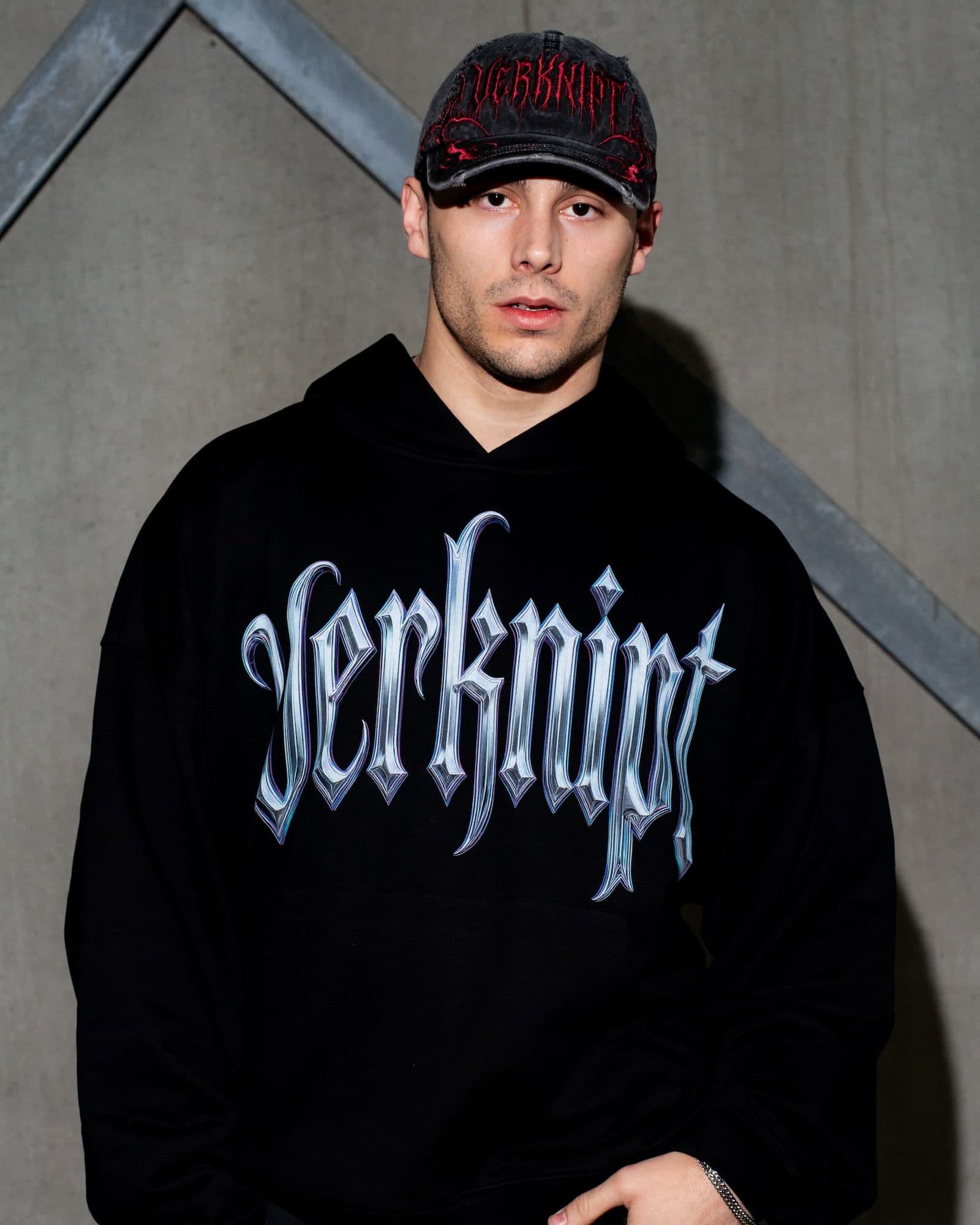 A person against a concrete wall with a diagonal steel beam, in a black hoodie with Verknipt in large chrome-look gothic letters across the chest and a washed black cap with VERKNIPT embroidered in red.