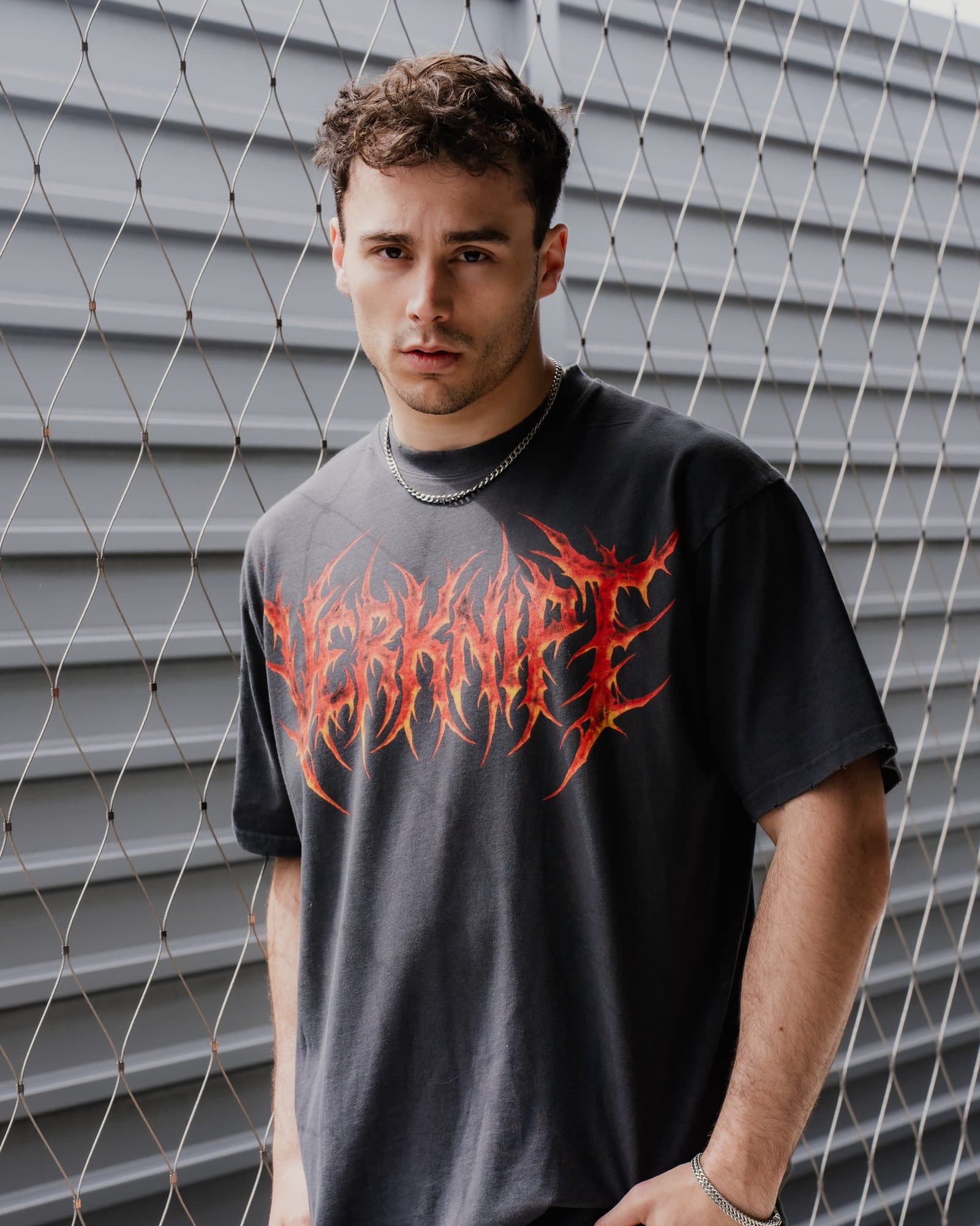 A person with curly dark hair in front of a steel mesh fence and a grey corrugated wall, in a washed black T-shirt with VERKNIPT in large red and orange flame letters across the chest and a silver chain.
