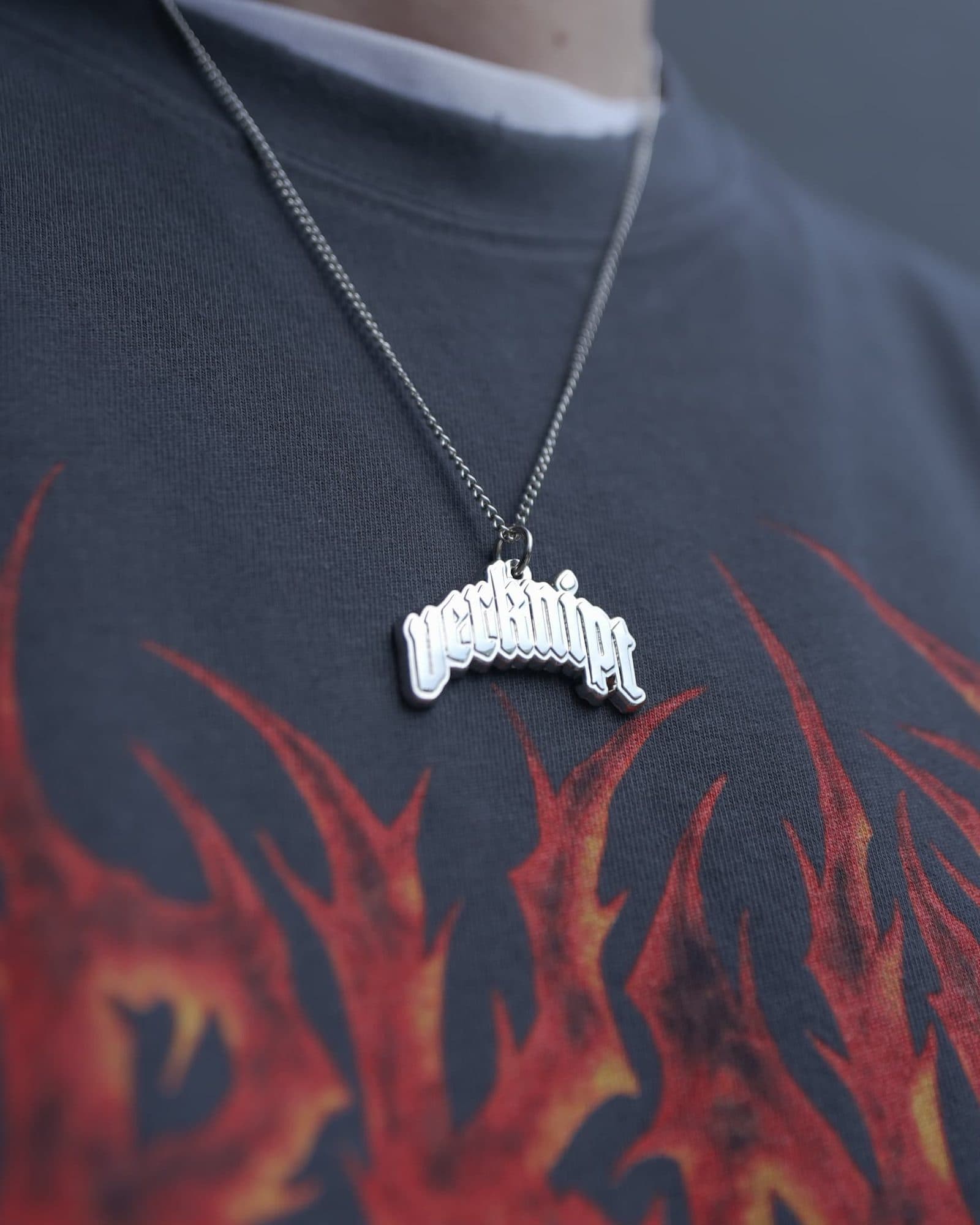 Close-up of a silver pendant in the shape of the verknipt wordmark on a fine chain, worn over a washed charcoal T-shirt with a large red and orange flame print below the collar.