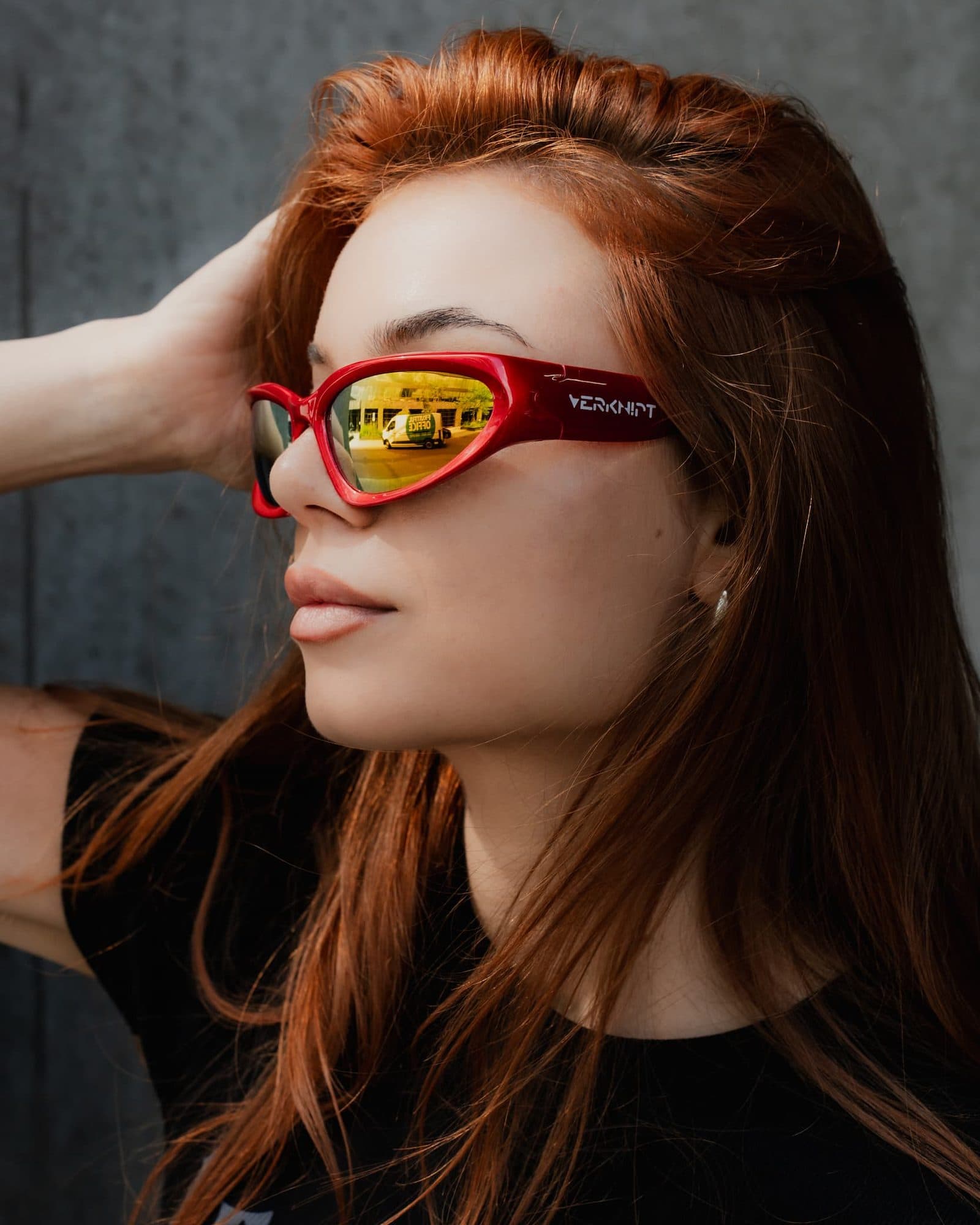Profile of a person with long red hair, one hand in the hair, in front of a dark grey wall, in glossy red wraparound sunglasses with mirrored gold lenses and VERKNIPT in white on the arm, over a black T-shirt with pale grey lettering partly hidden by the hair.