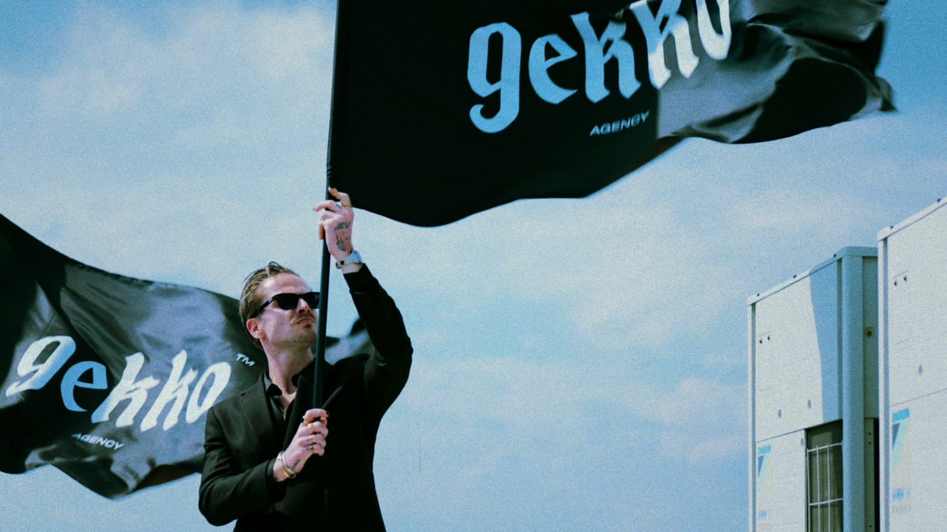 Still from the GEKKO Agency film: a person in sunglasses holding up a large black GEKKO Agency flag on a roof under a blue sky, a second flag beside them