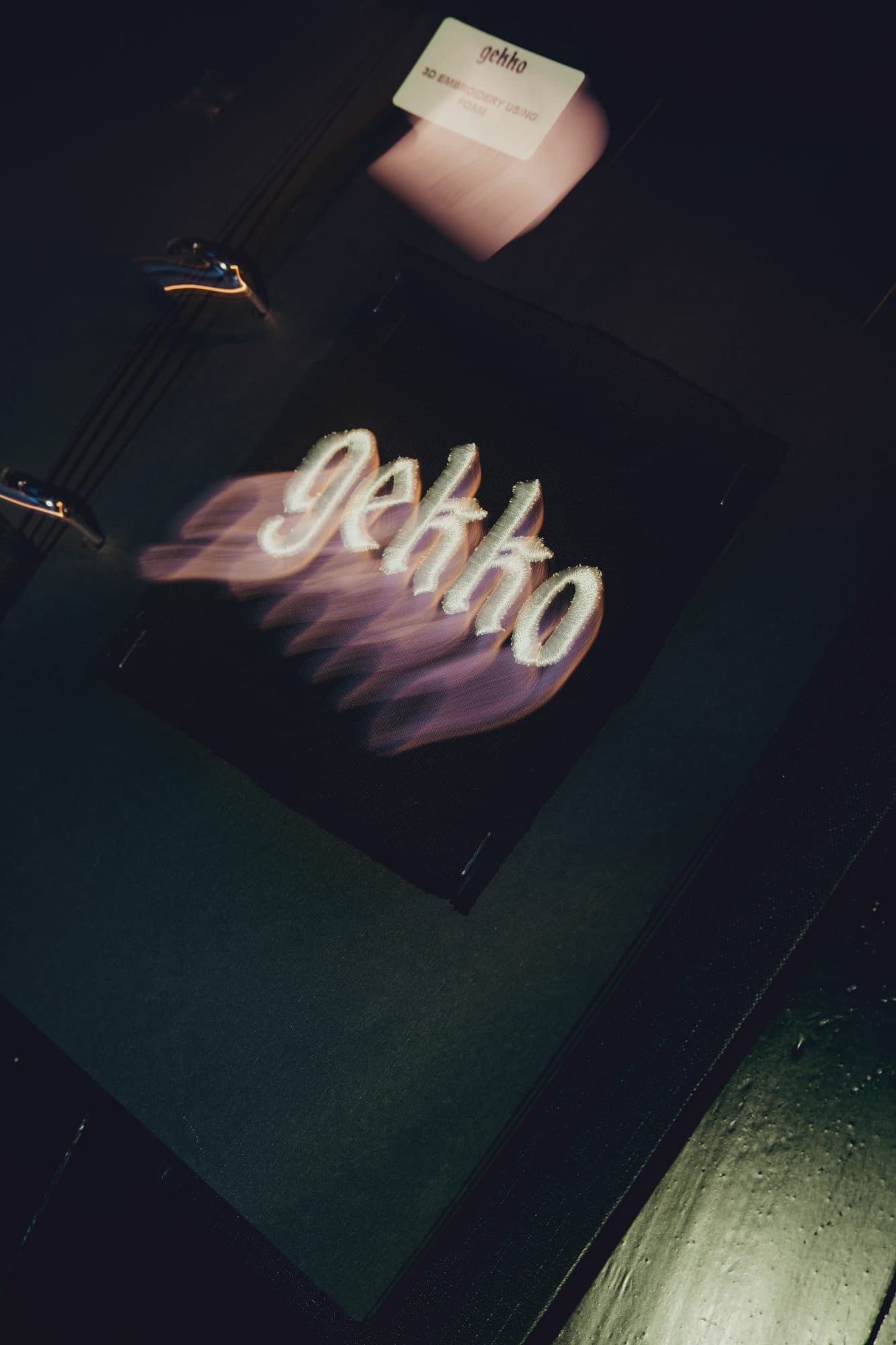 Raised white foam GEKKO embroidery on black fabric with light trails from a long exposure, below a technique label.